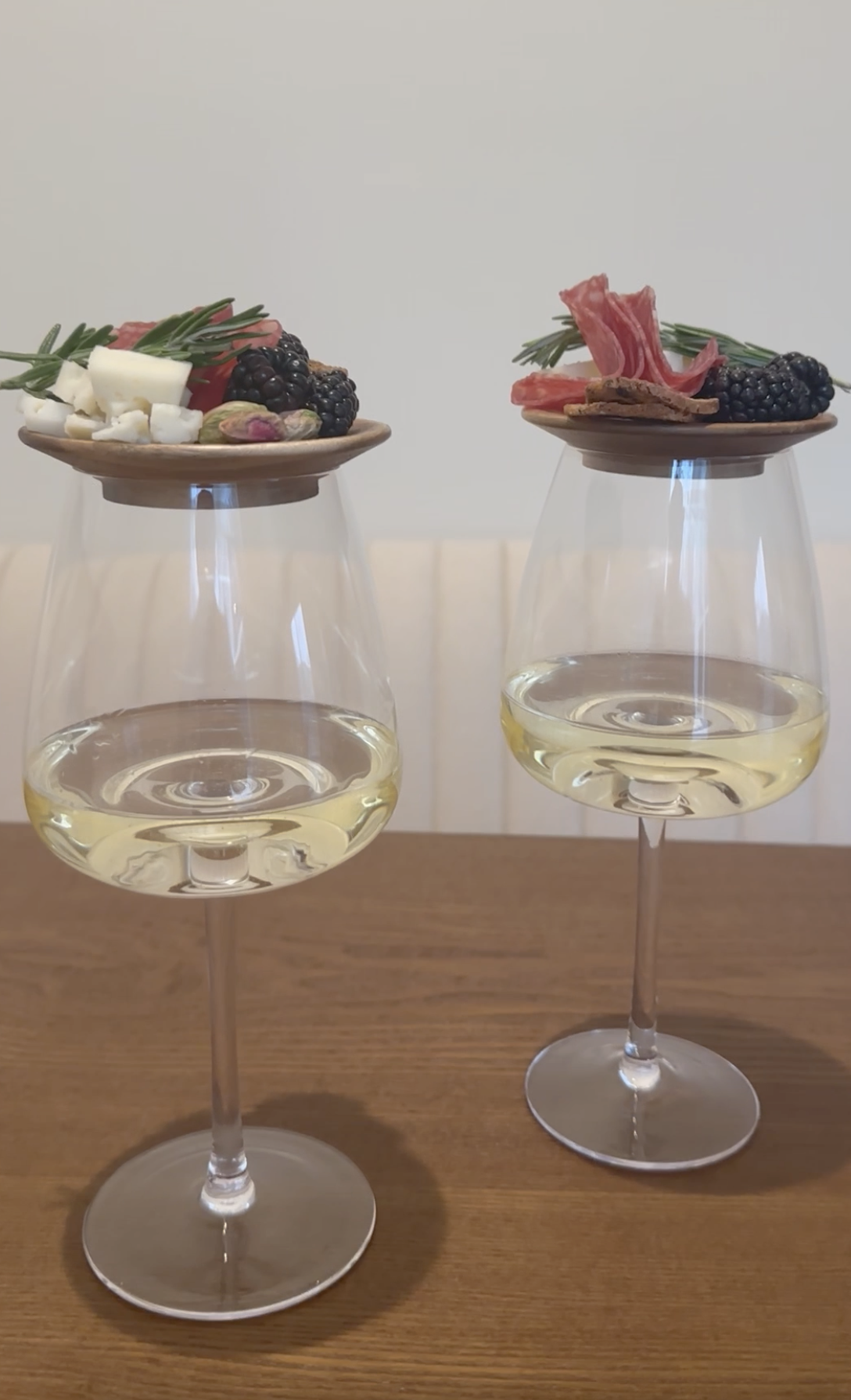Two wine glasses filled with white wine, each balanced on a small wooden plate with cheese, blackberries, slices of meat, and herbs.