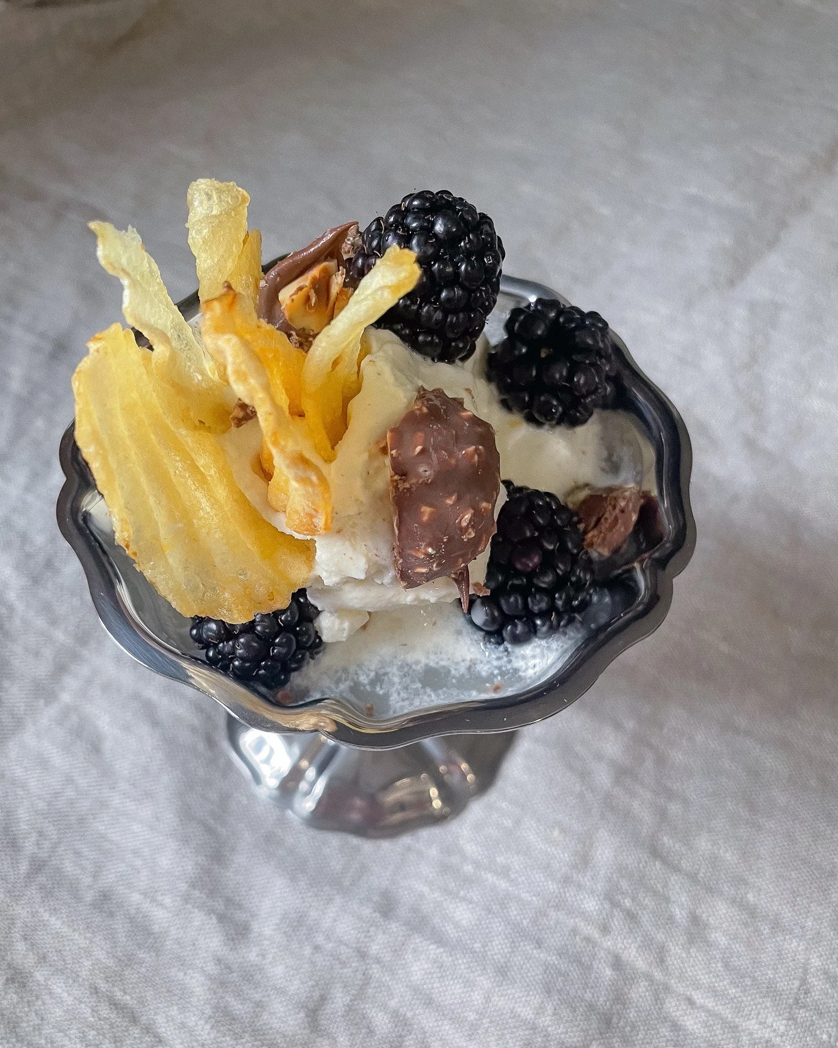 A dessert cup filled with ice cream, topped with blackberries, a piece of chocolate, and crispy cheese or cookie strips.