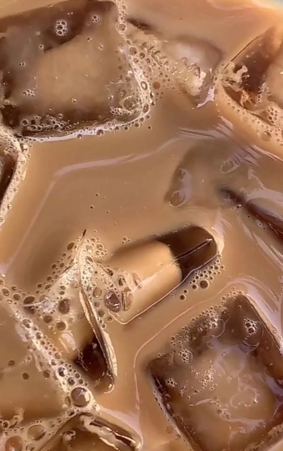 Close-up of ice cubes in a container of coffee with milk, showing bubbles and swirls.
