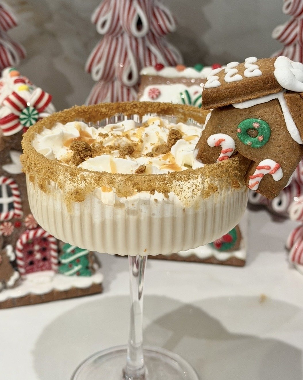A dessert cocktail glass filled with a creamy beverage topped with whipped cream, caramel drizzle, and crumbled cookies. A decorated gingerbread house cookie is attached to the rim of the glass. The background features festive holiday candy and cookie decorations.
