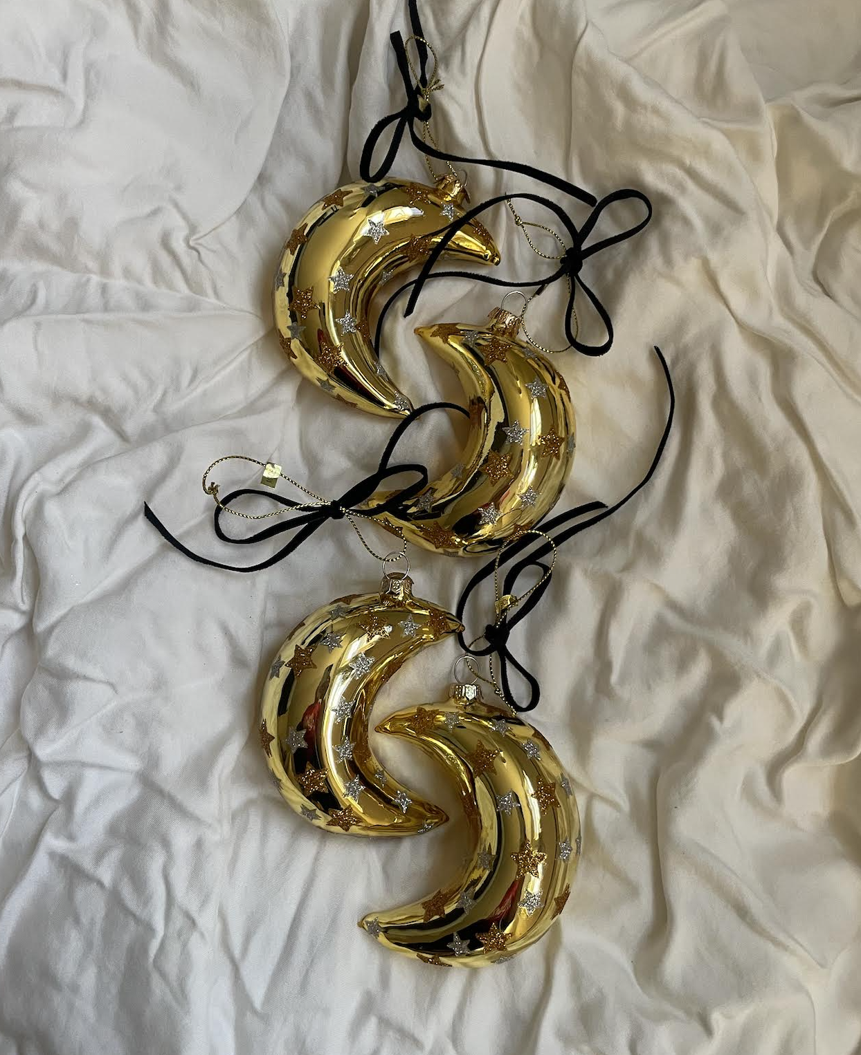 Four gold crescent moon-shaped Christmas ornaments with star decorations, hanging on black ribbons, laid on white crumpled fabric.