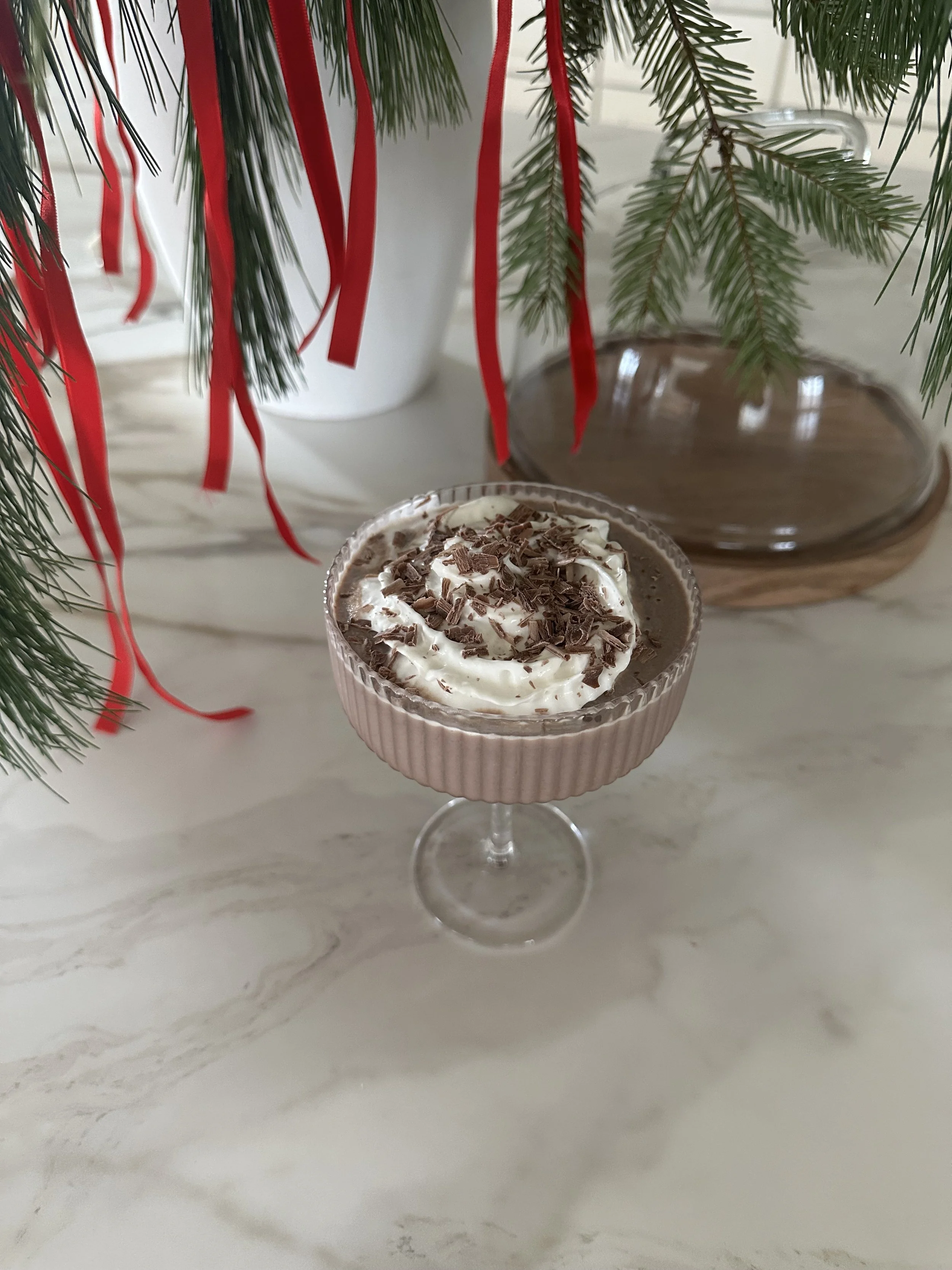 A pink cocktail glass with whipped cream and chocolate shavings on top, placed on a white marble surface. Green pine branches with red ribbons are hanging above, and a wooden tray is in the background.