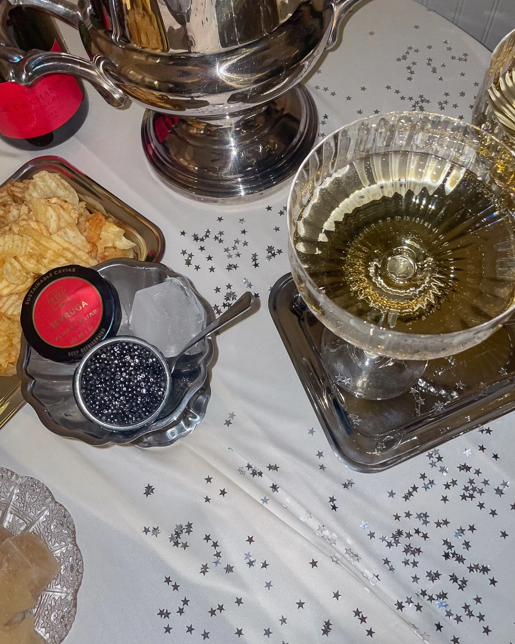 A celebration table setup with a silver trophy, a glass of white wine, a bowl of caviar with a spoon, a plate of chips, and holiday-themed decorations with star-shaped confetti.