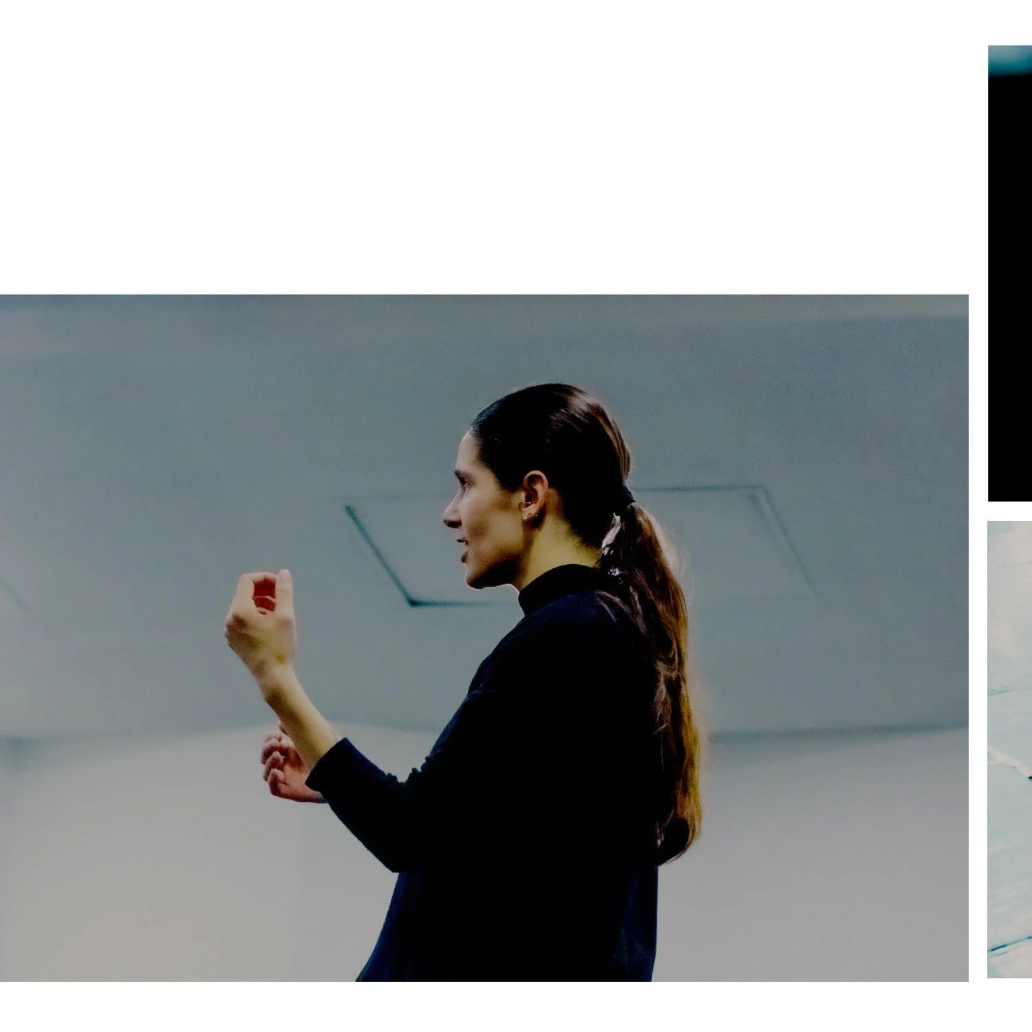 Side view of a woman with long dark hair tied back, wearing a black top, gesturing with her right hand while speaking in a modern indoor setting.