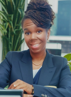 Smiling woman in a blue blazer sitting at a desk in an office with green plants in the background.