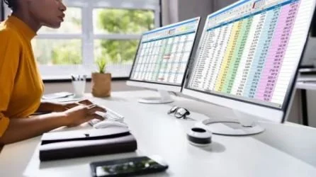 Person working at a desk with two computer monitors displaying spreadsheets.