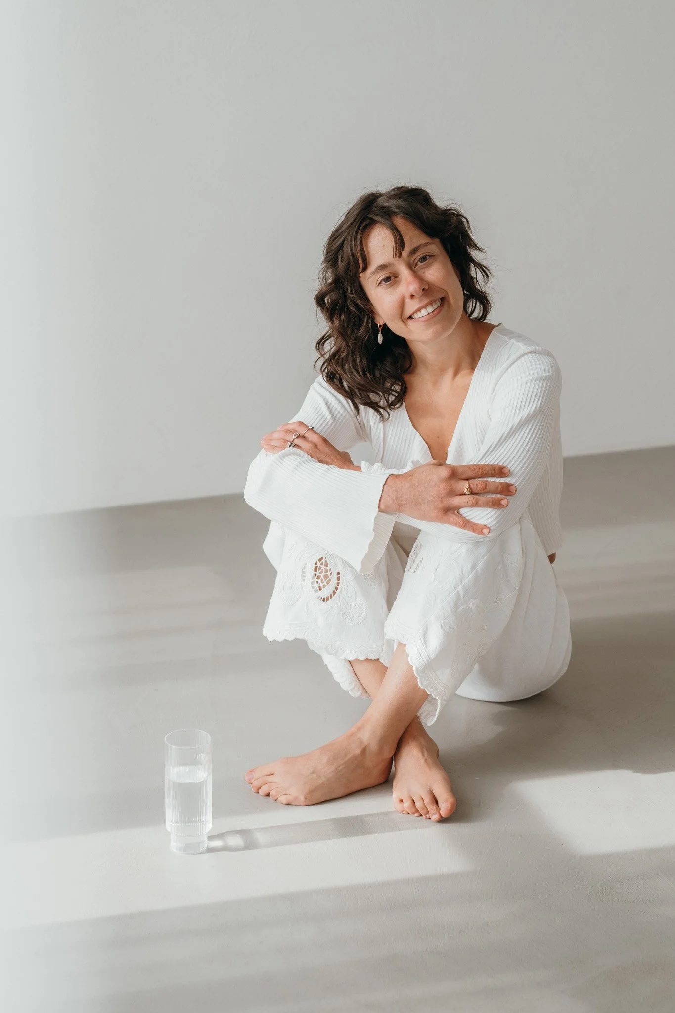 A woman with curly brown hair smiling while sitting on the floor in a white yoga outfit, with a glass of water nearby, in a minimalistic room. Photo by theneutralstudio.ch