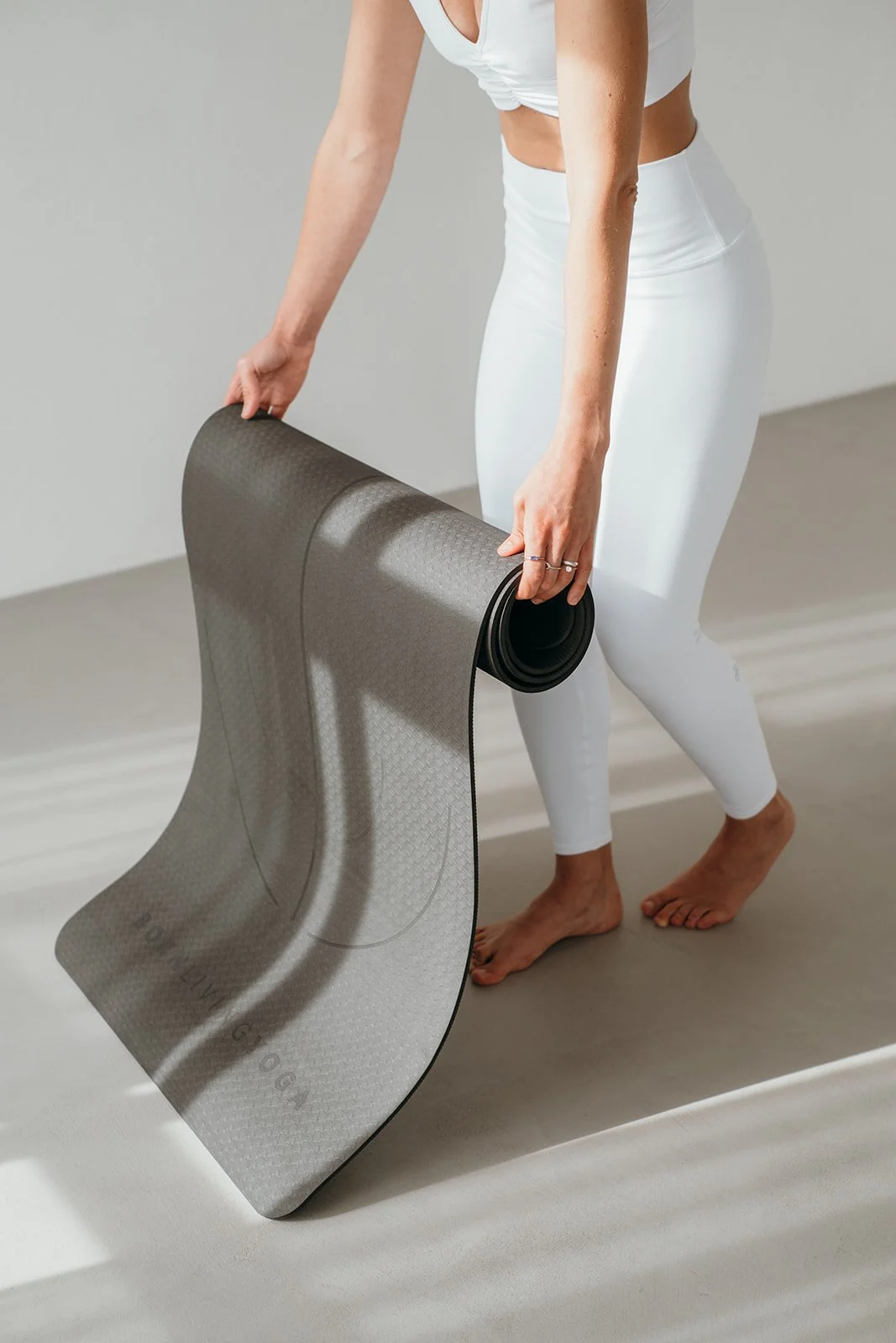 Person in white athletic wear unrolling a gray yoga mat on a neutral-colored floor.