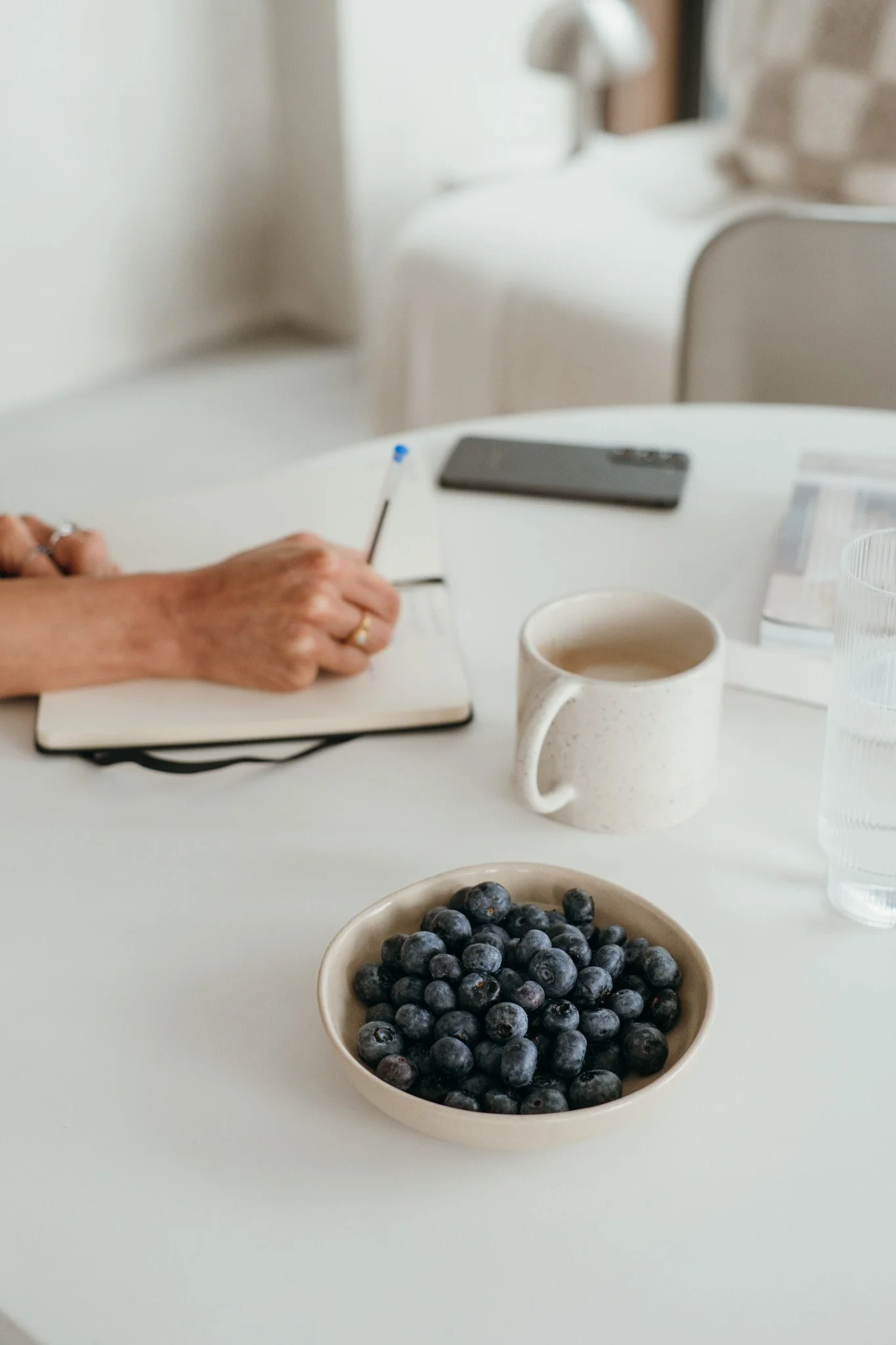A white table with a bowl of blueberries, a ceramic mug with a beverage, a glass of water, a smartphone, a notebook with a person writing, and a magazine or booklet.