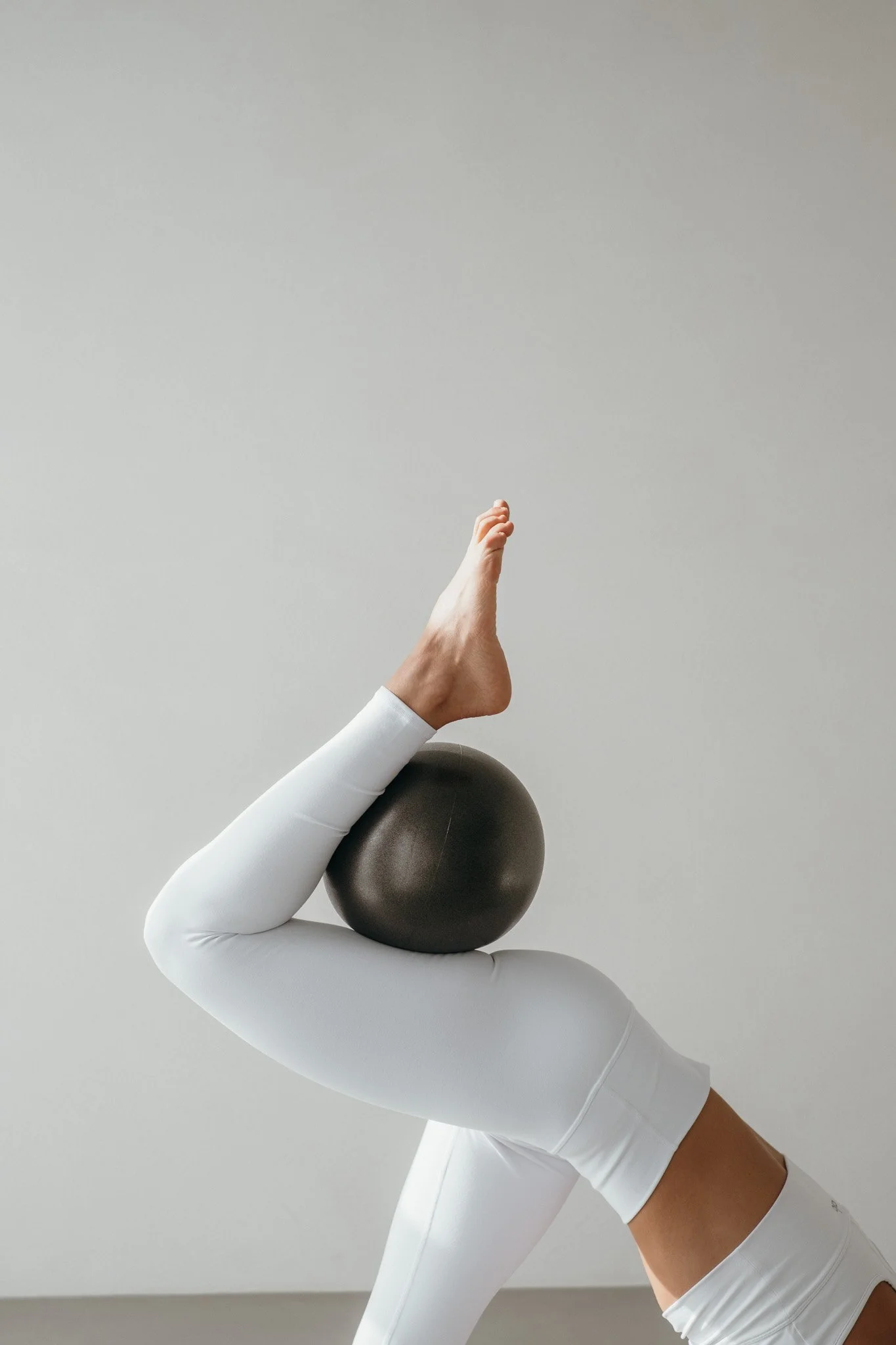 A person in white workout clothes balancing a black pilates ball with their leg, with their foot pointed upward.