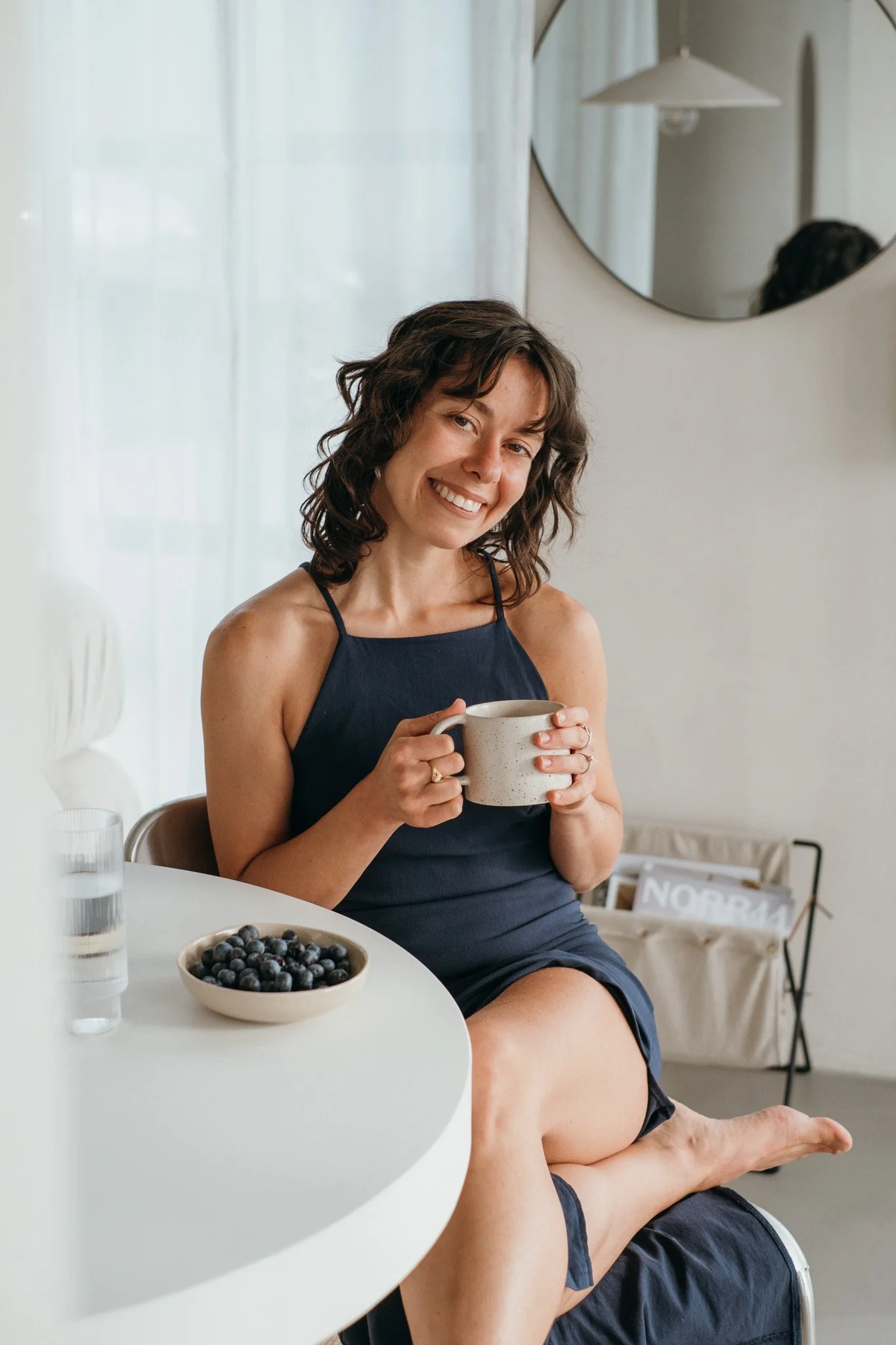 A woman sitting at a table, smiling and holding a mug, with a bowl of blueberries and a glass of water in front of her, in a bright, modern kitchen. Photo by theneutralstudio.ch