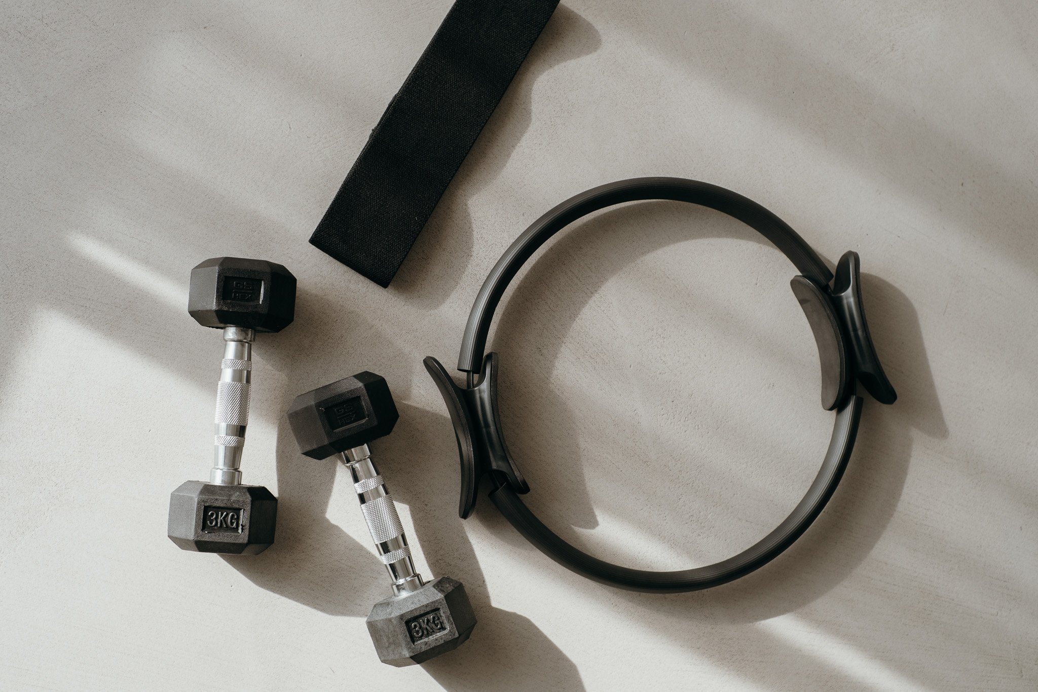 Pilates equipment including two 3 kg dumbbells, a resistance band, and a pilates ring on a light-colored surface. Photo by theneutralstudio.ch