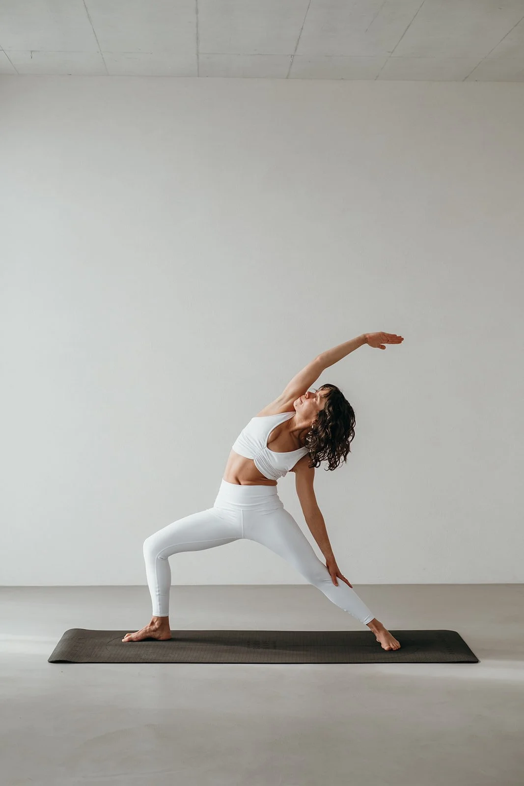 Woman practicing yoga in a wide stance on a dark grey yoga mat, stretching with one arm overhead and the other reaching toward her ankle in a bright, minimal room.