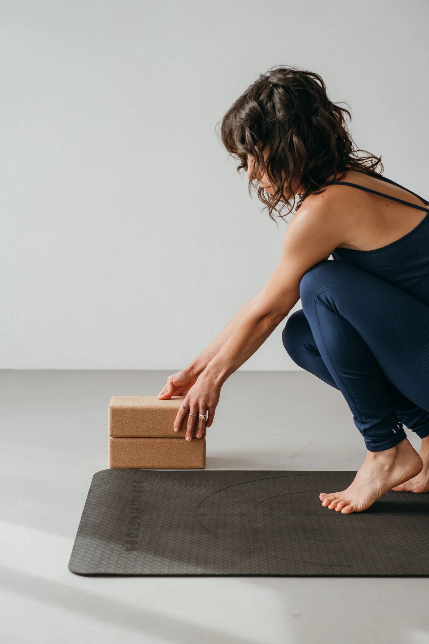 A woman in indigo workout clothing adjusting a yoga block next to her yoga mat.