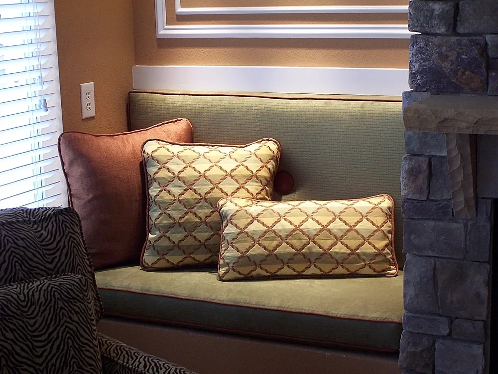 A cozy seating area with a built-in bench, three decorative cushions, a stone fireplace, a window with blinds, and a framed picture on the wall.