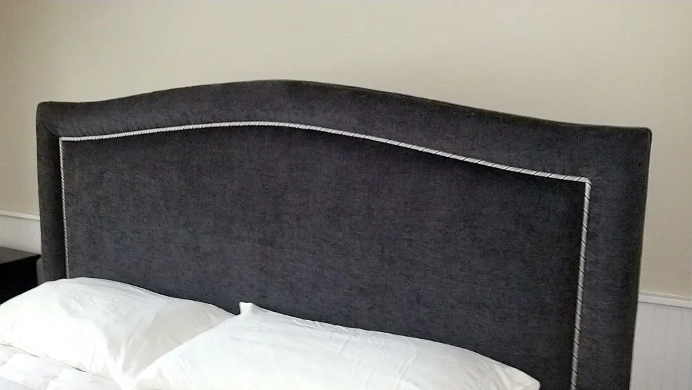 A black upholstered bed headboard with white piping, placed against a beige wall, with white pillows and bedding.