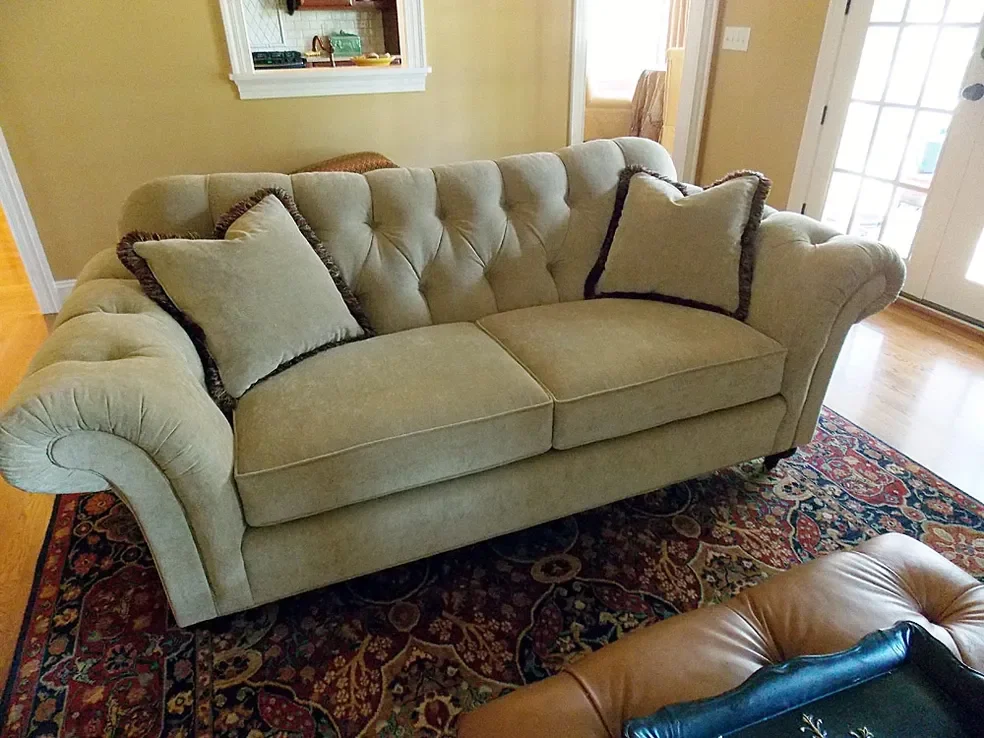 A beige tufted sofa with two matching pillows on a colorful Persian-style area rug inside a living room.