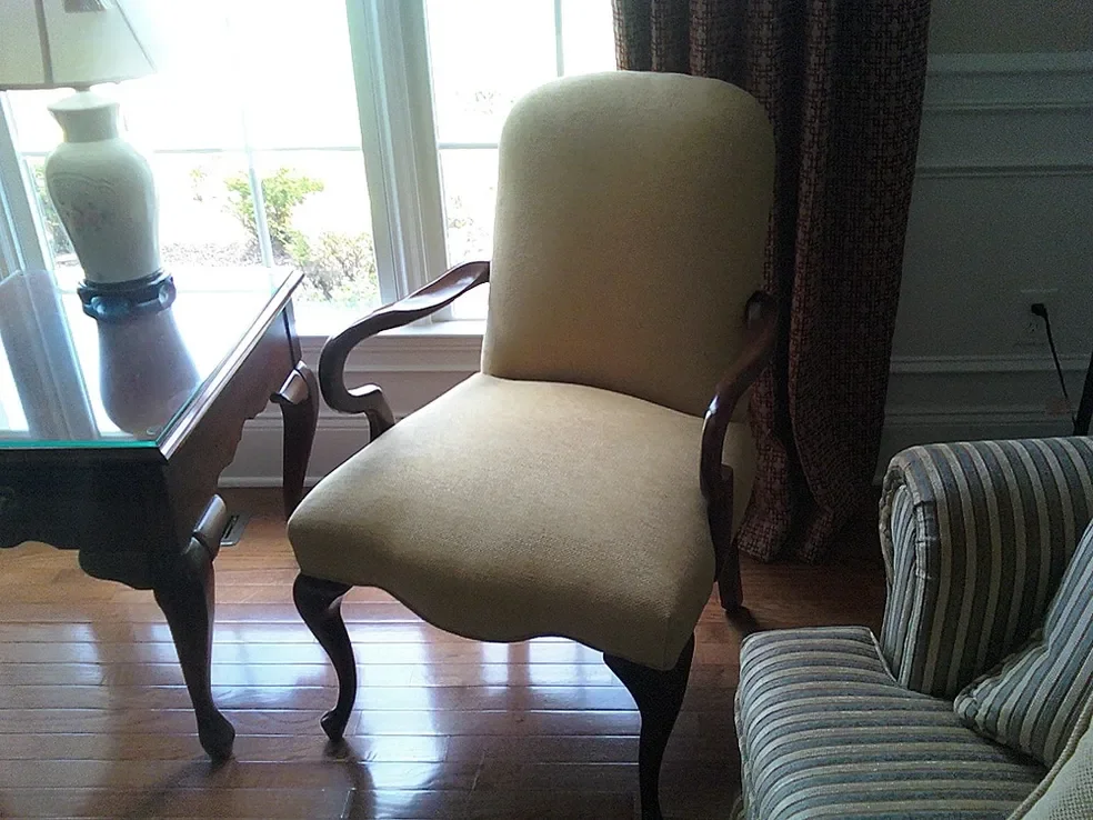 A beige upholstered armchair with dark wooden legs and armrests, placed next to a striped armchair with a pillow, near a window with curtains and a glass-top wooden table with a white vase, in a lit room with hardwood flooring.