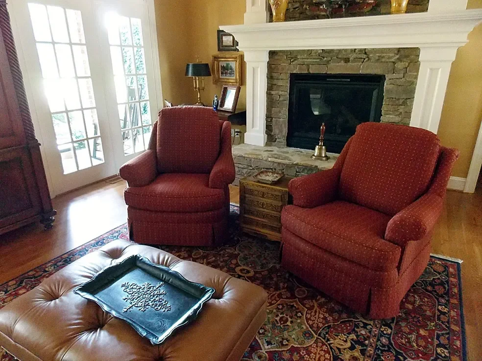 Living room with two red upholstered armchairs, a leather ottoman with a decorative tray, and a brick fireplace with a mantle. There is a wooden side table with a brass bell, a lamp, and picture frames. French doors lead outside, and a patterned rug