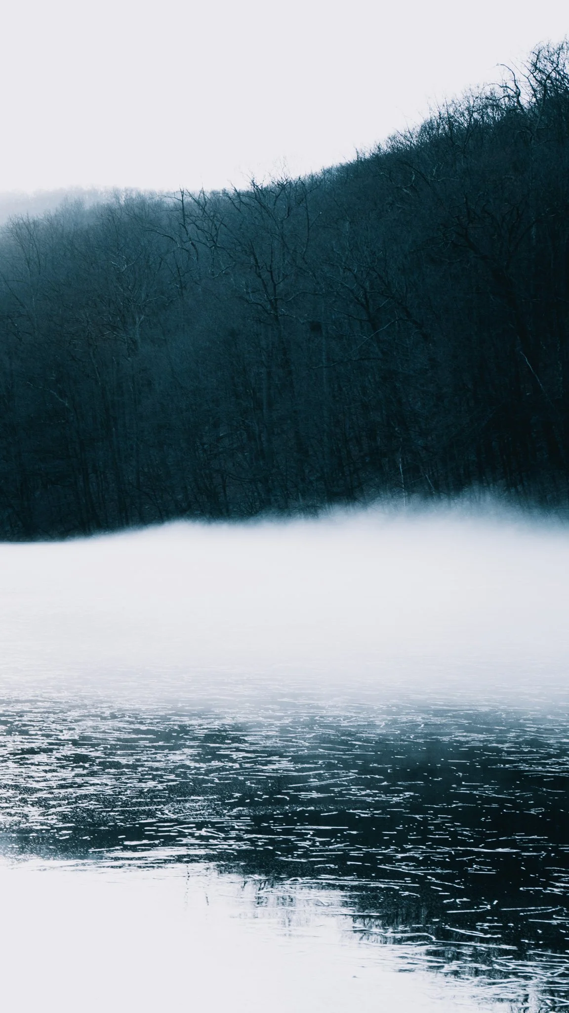 A winter landscape of a lake with fog, reflections on the water, and a forested hill in the background. Matching with its twin photo, the wooden dock scene.