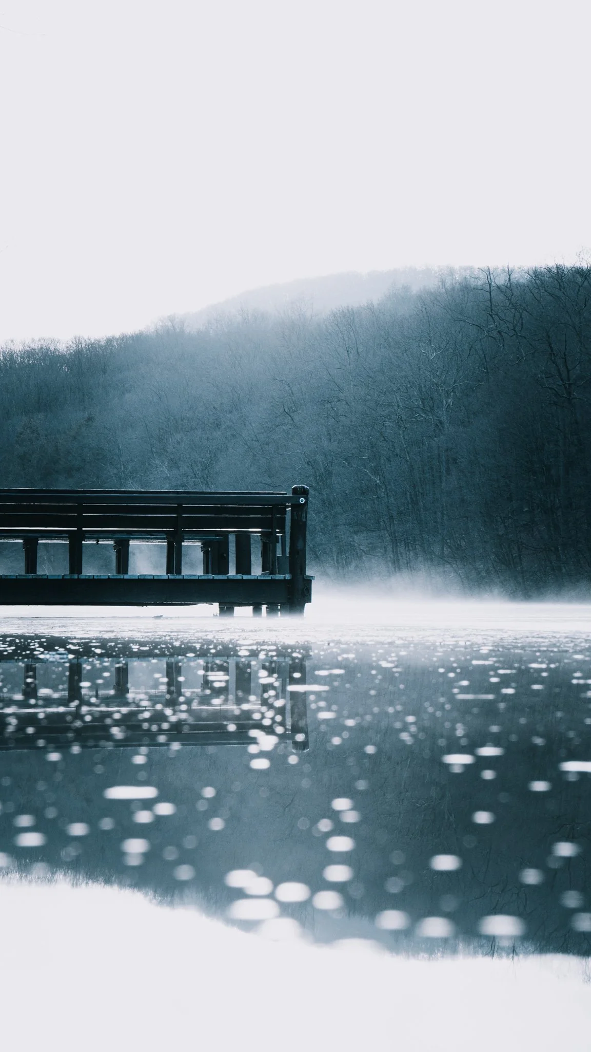 A winter scene showing a wooden dock extending over a partially frozen lake, with a backdrop of snow-covered trees and foggy mountains. The water has reflections and bokeh lights.
