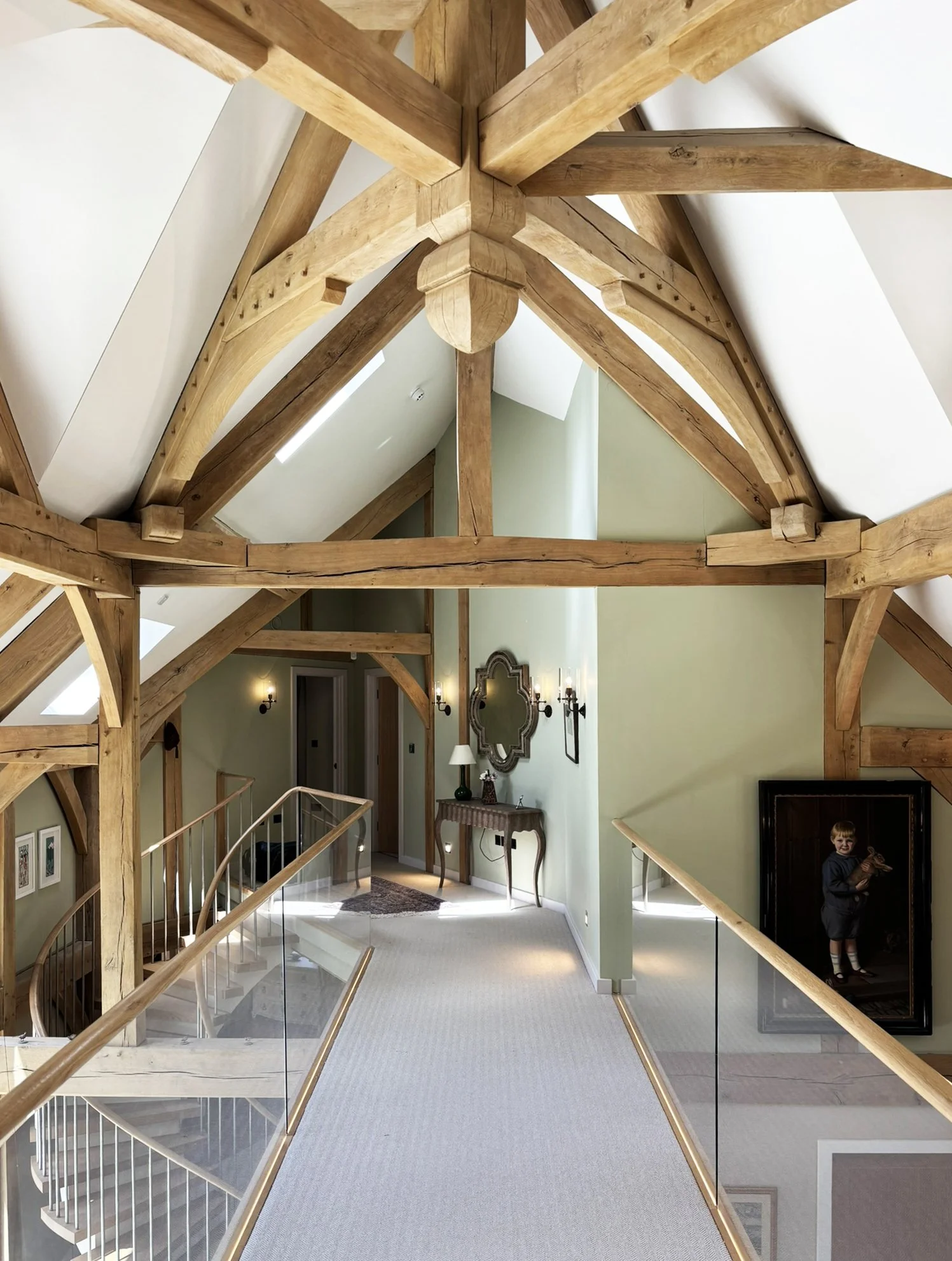 Sarsen Architects — View of a spacious, lit attic with exposed wooden beams, a high ceiling, a decorative mirror, a small table with a lamp, and a young boy standing in a