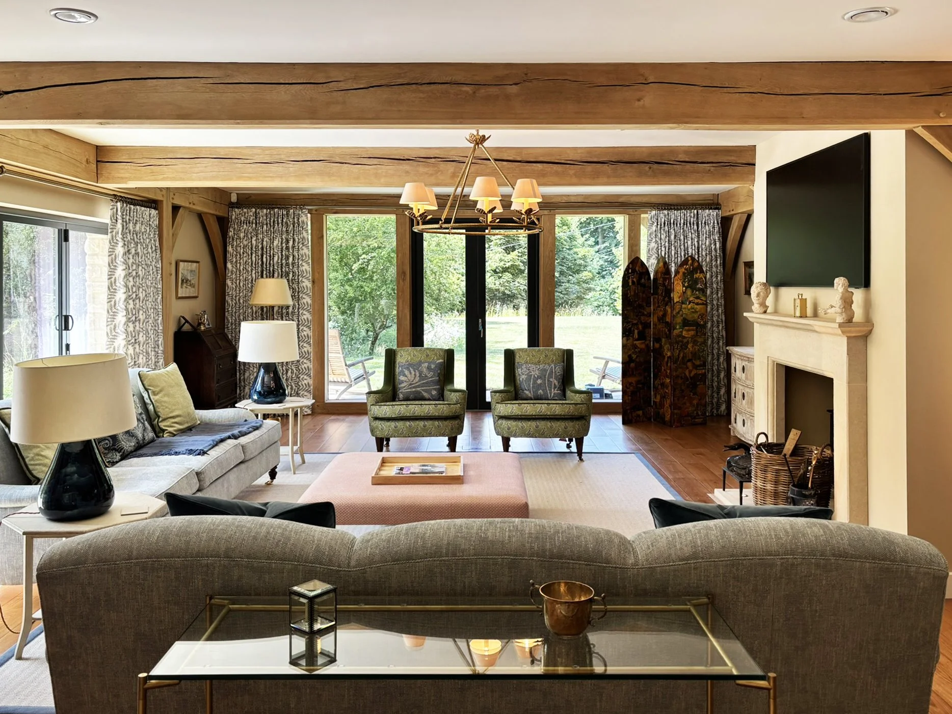 Sarsen Architects — Living room with beige couch, green armchairs, wooden beams, sliding glass door, and fireplace, overlooking a garden
