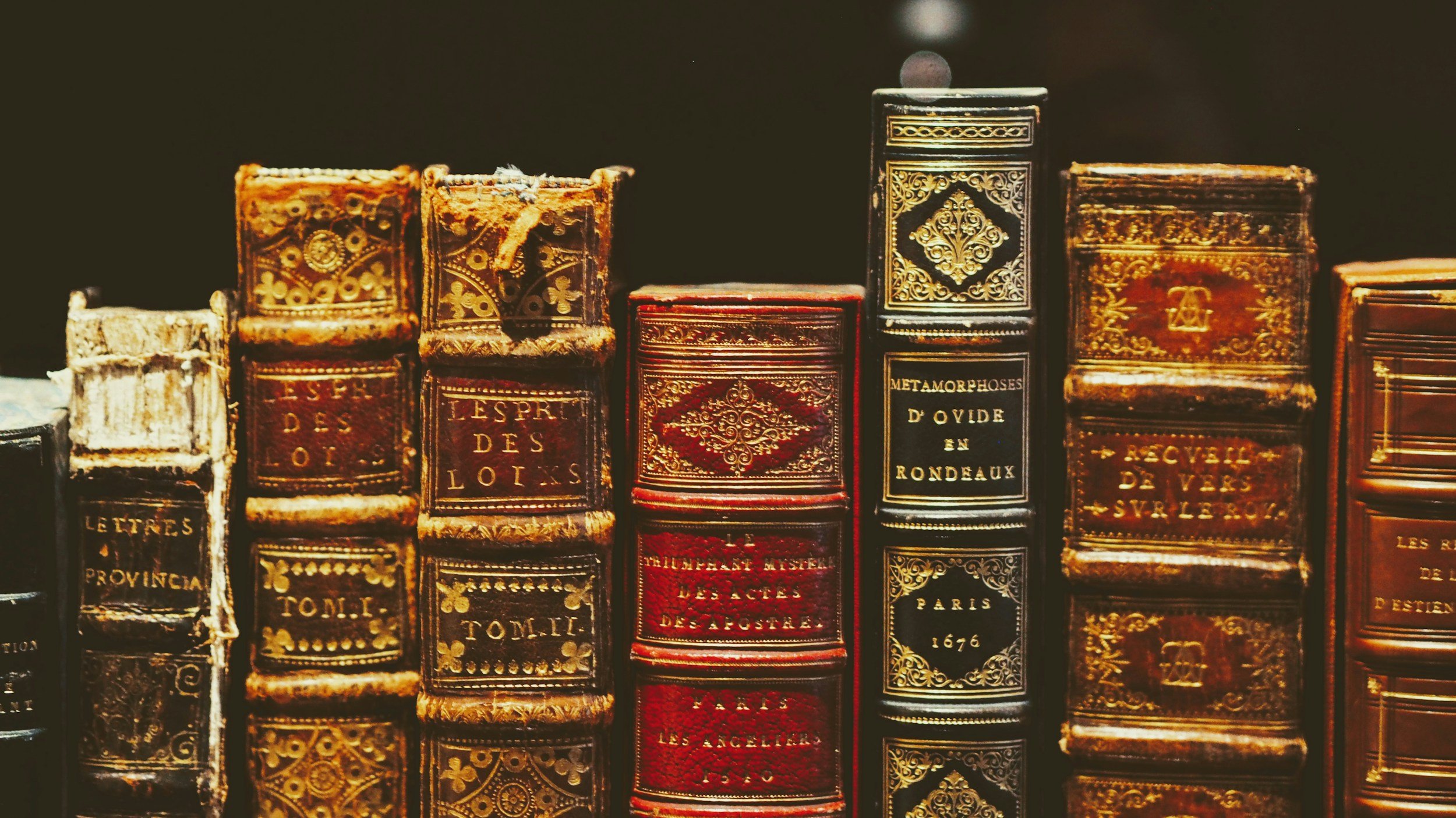 A row of vintage, ornate hardcover books with gold embossing on their spines, some with worn covers, laid on a shelf.