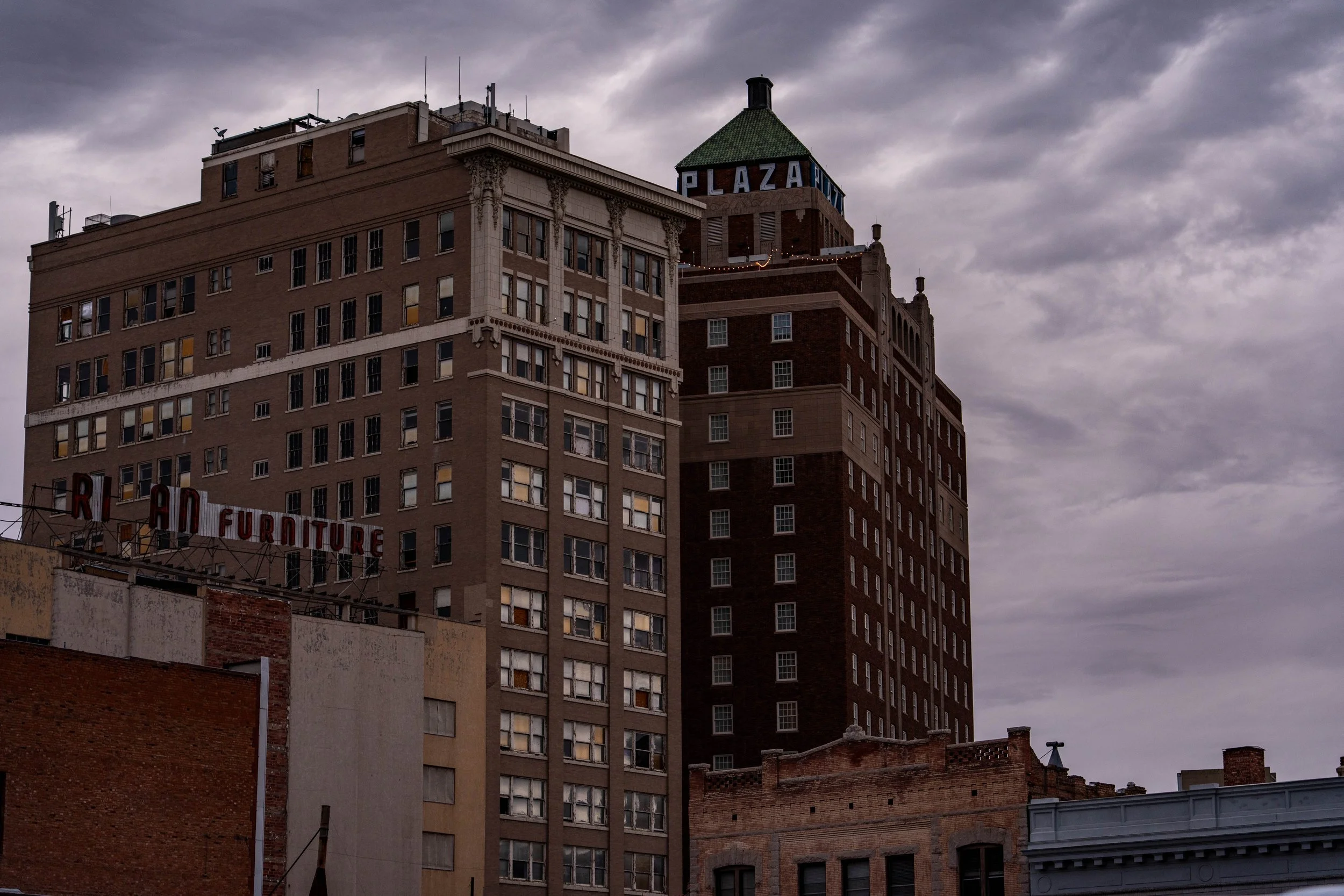 Cityscape with multiple tall buildings under an overcast sky, including a brown brick building with a green-roofed tower and a sign that says 'PLAZA' on top.