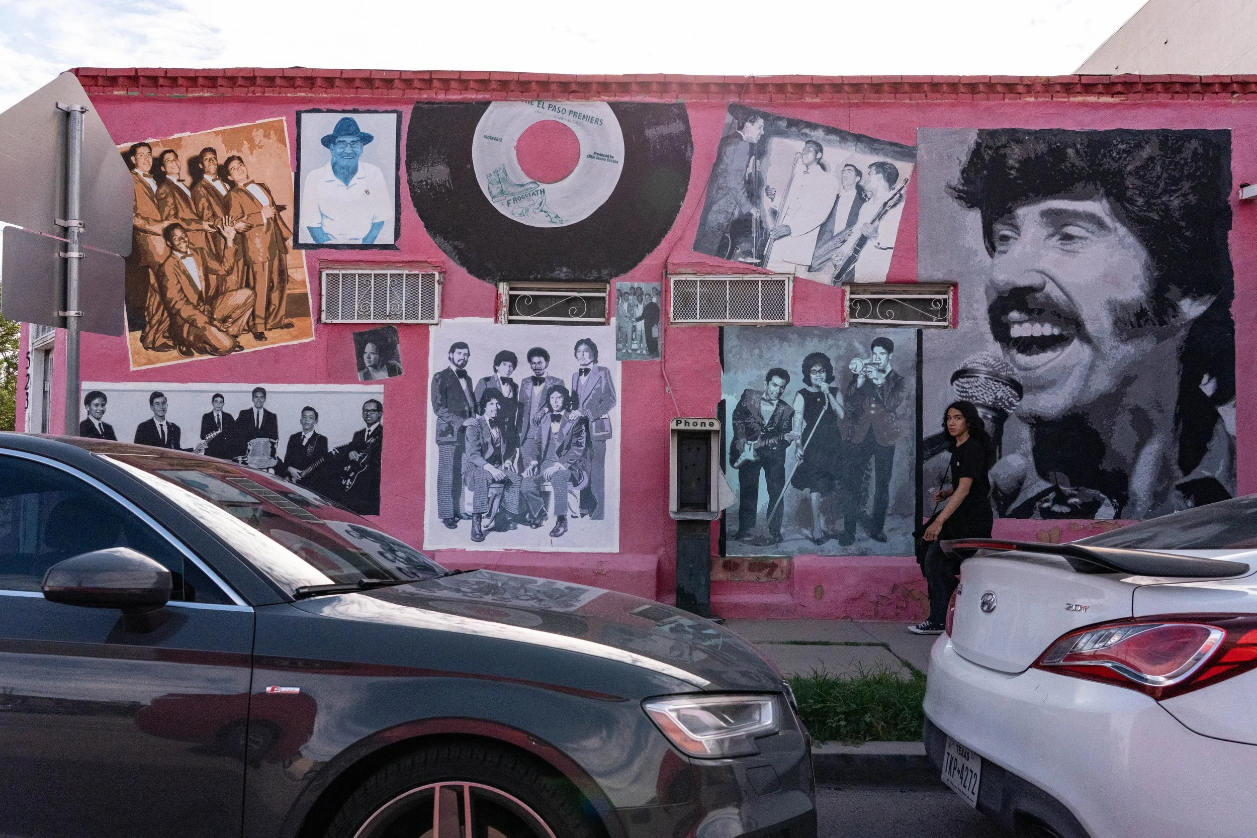 Mural on pink wall depicting various black and white images of musicians, singers, and performers, including a large portrait of a woman singing into a microphone, with cars parked in front and a woman in black standing nearby.