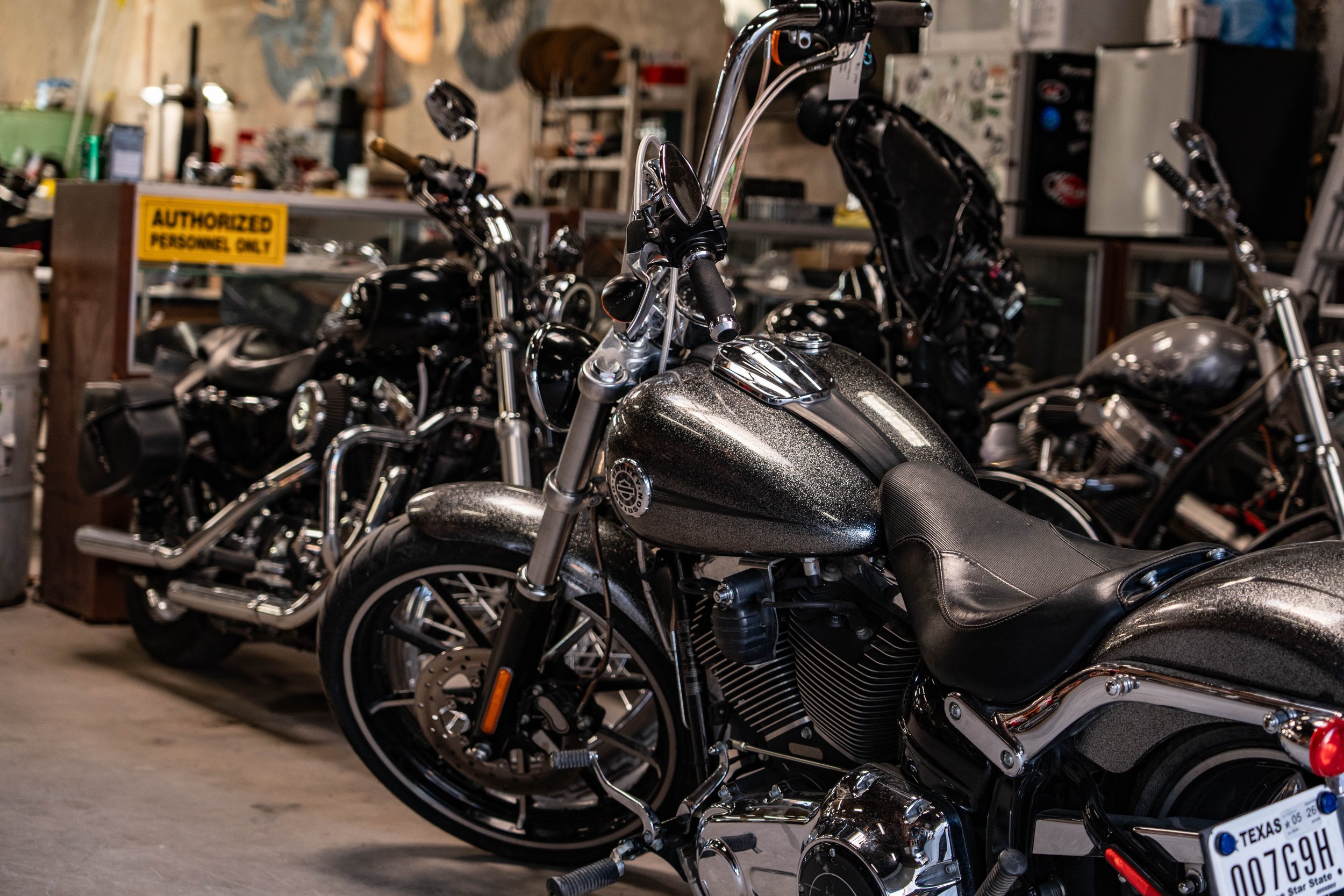 A row of black motorcycles parked inside a garage or workshop with a sign that reads 'Authorized Personnel Only' in the background.