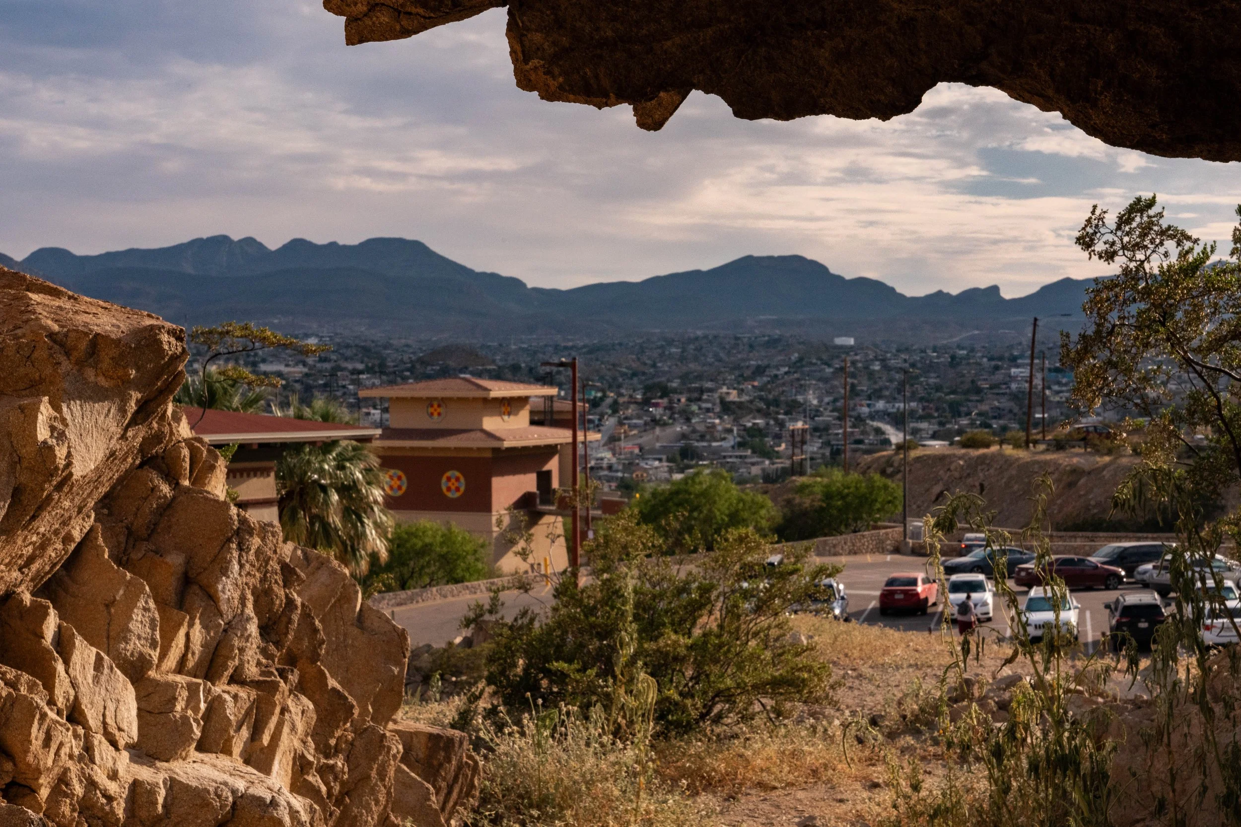 View of a cityscape with mountains in the background, framed by rocks and trees in the foreground, including a parking lot with cars.