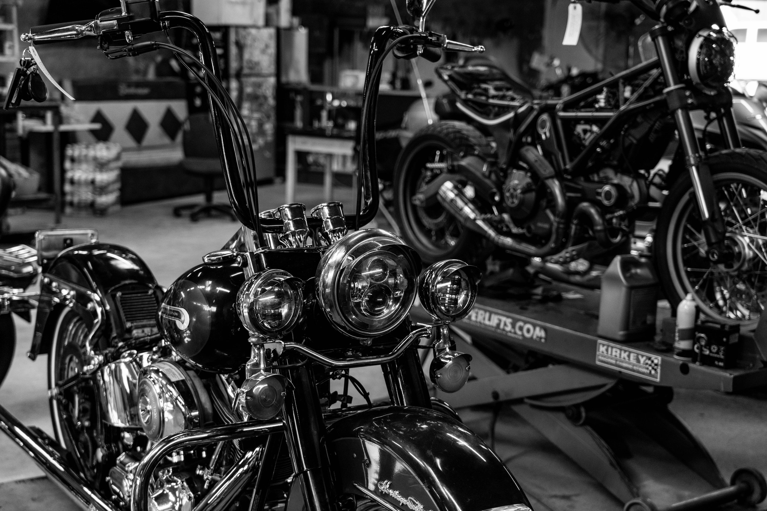 A black and white photo of a motorcycle in a garage, with another motorcycle on a trailer in the background.