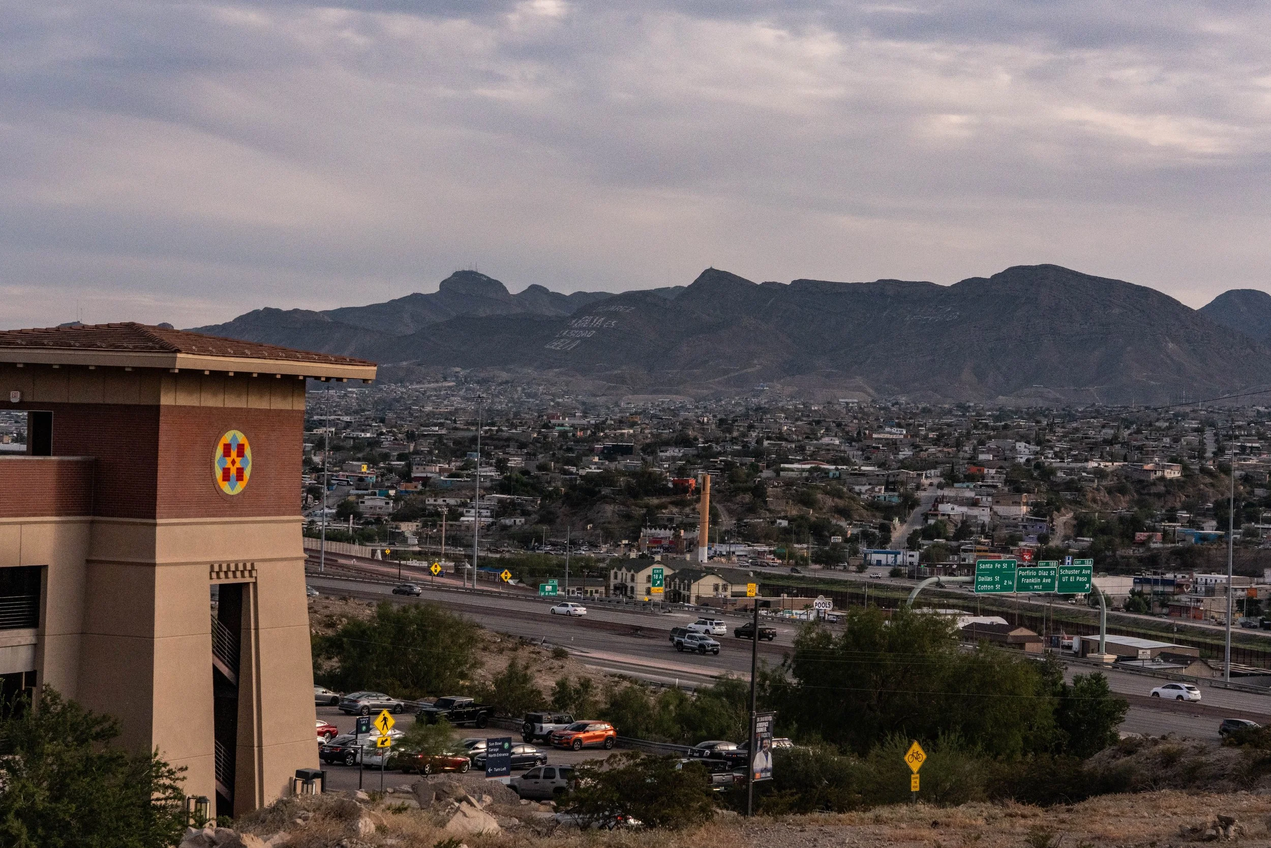 A cityscape with mountainous terrain in the background, highway with moving cars, and a building with a colorful emblem on its wall in the foreground.
