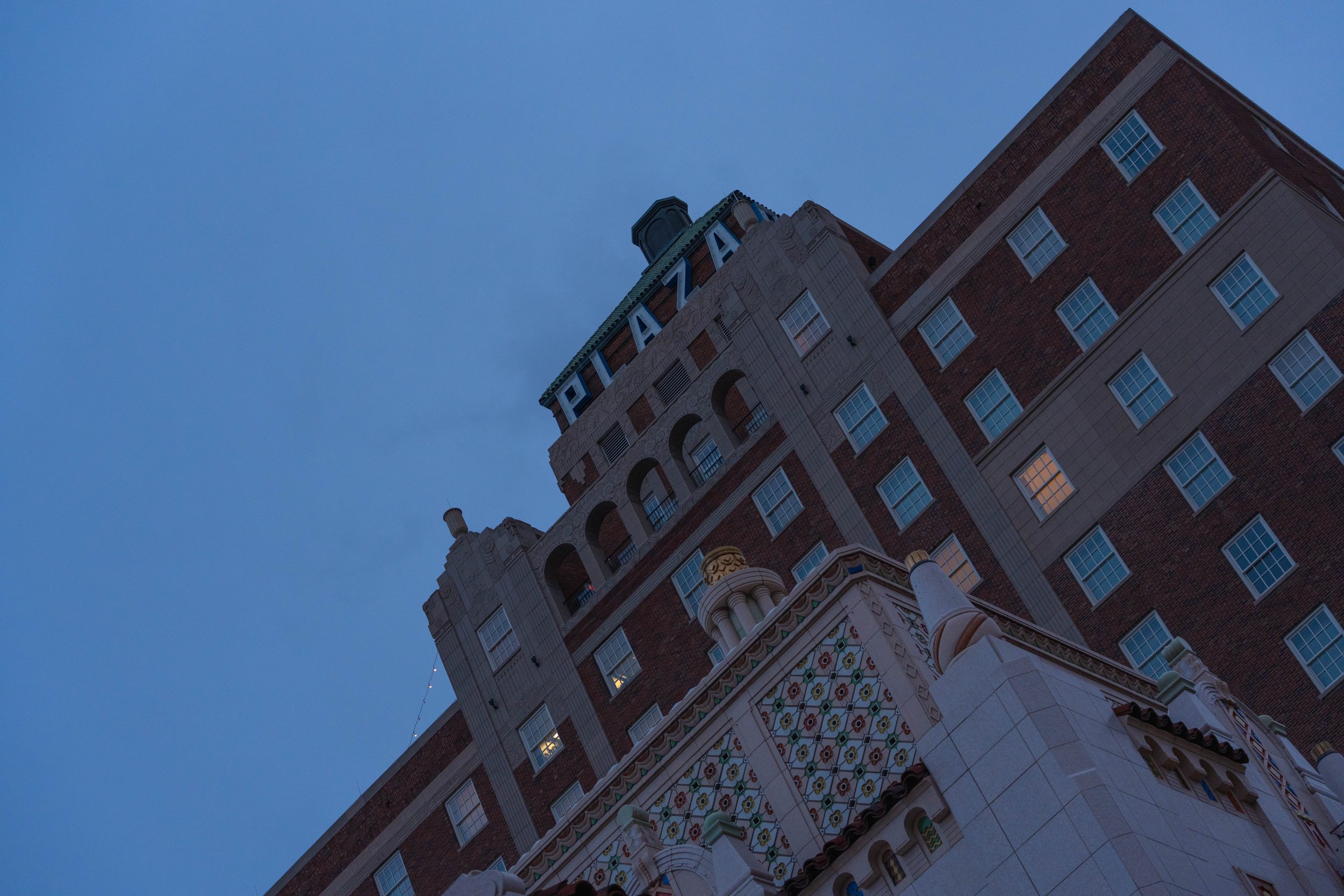 A tall building with a sign that reads 'PALAZA' on top, featuring arched windows and ornate architectural details, seen from a low angle against a blue sky.