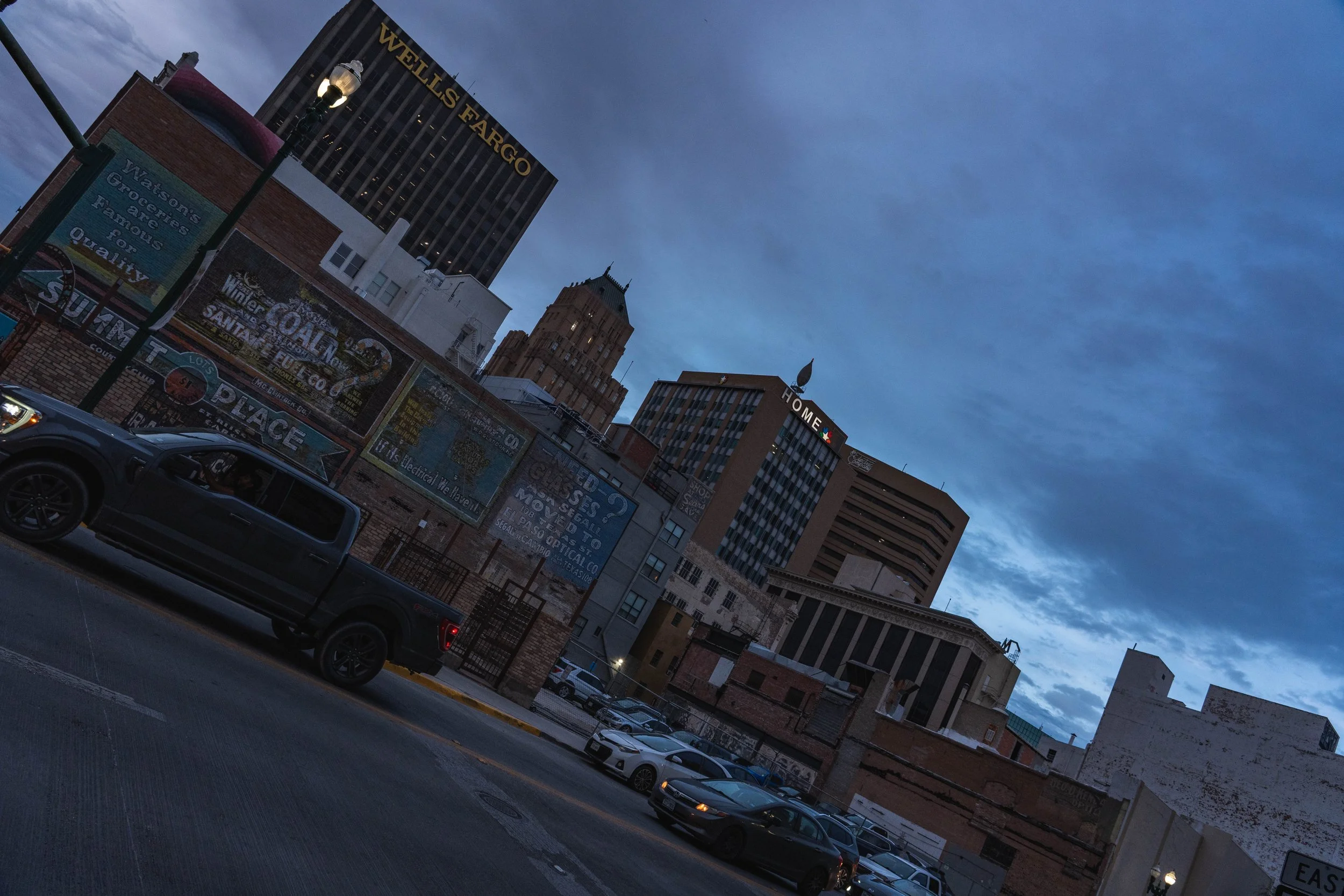 City street scene at dusk with tall buildings, including Wells Fargo and Home signs, parked cars, streetlamp, and cloudy sky.