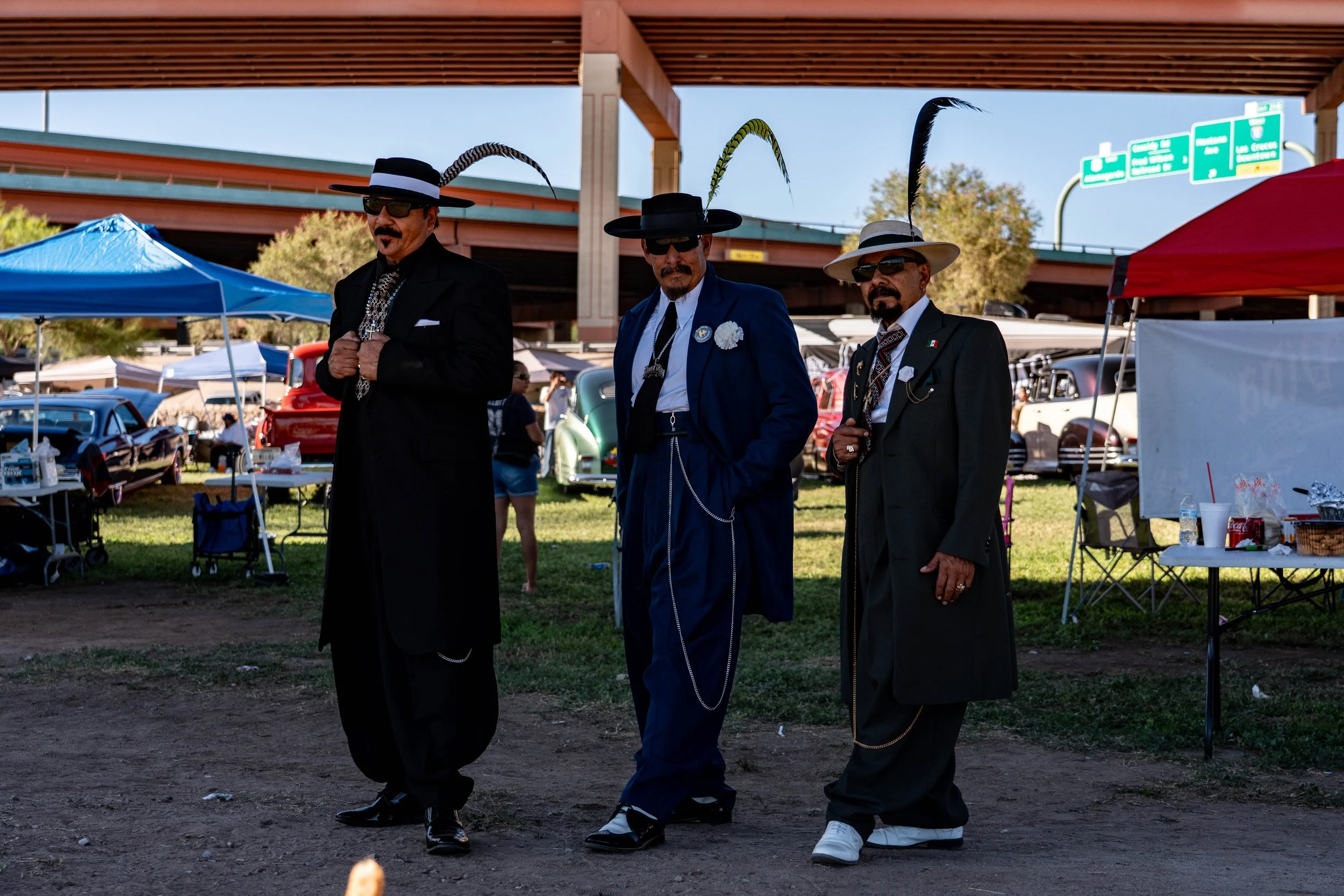 Three men dressed as Mexican drug lords, wearing black suits with chains and feathered hats, walking at an outdoor car show.