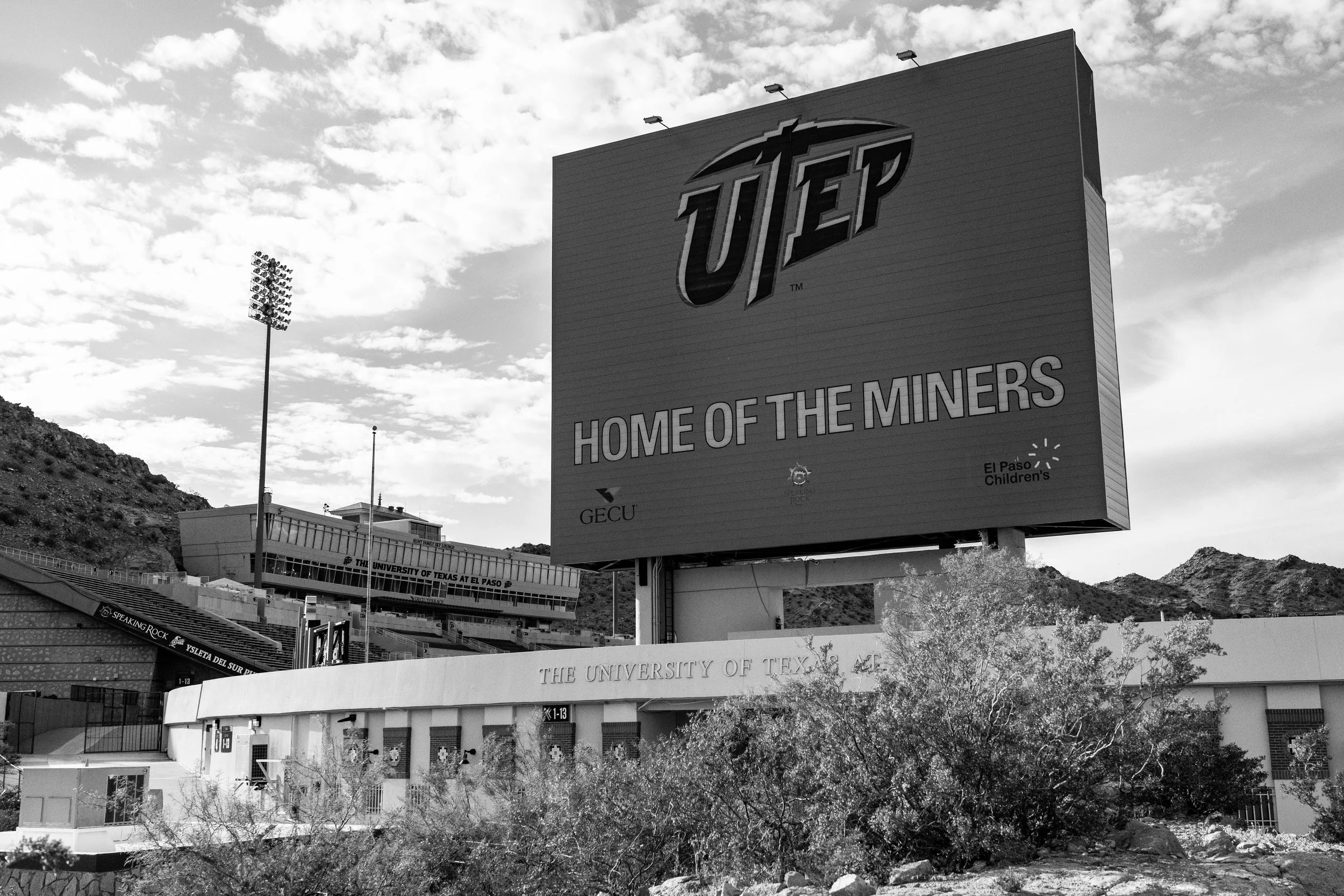 A large billboard at The University of Texas at El Paso featuring the UTEP logo and the slogan "Home of the Miners" with surrounding buildings, hills, and a cloudy sky in the background.