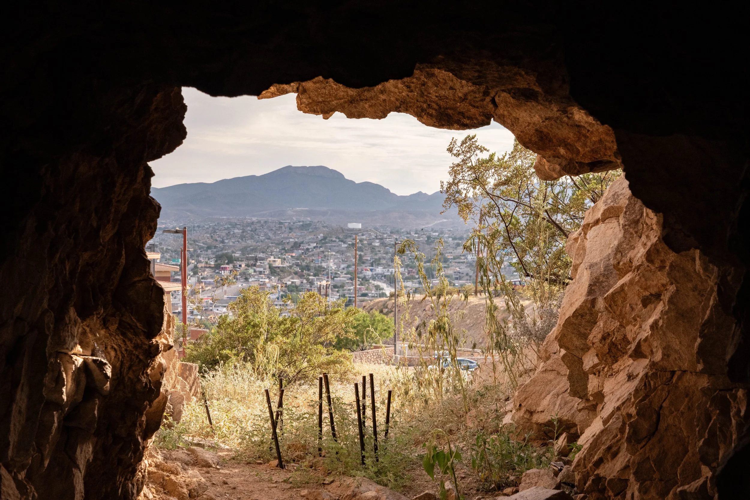 View of a city with mountain in the background, seen through a rocky cave opening, with desert plants in the foreground.