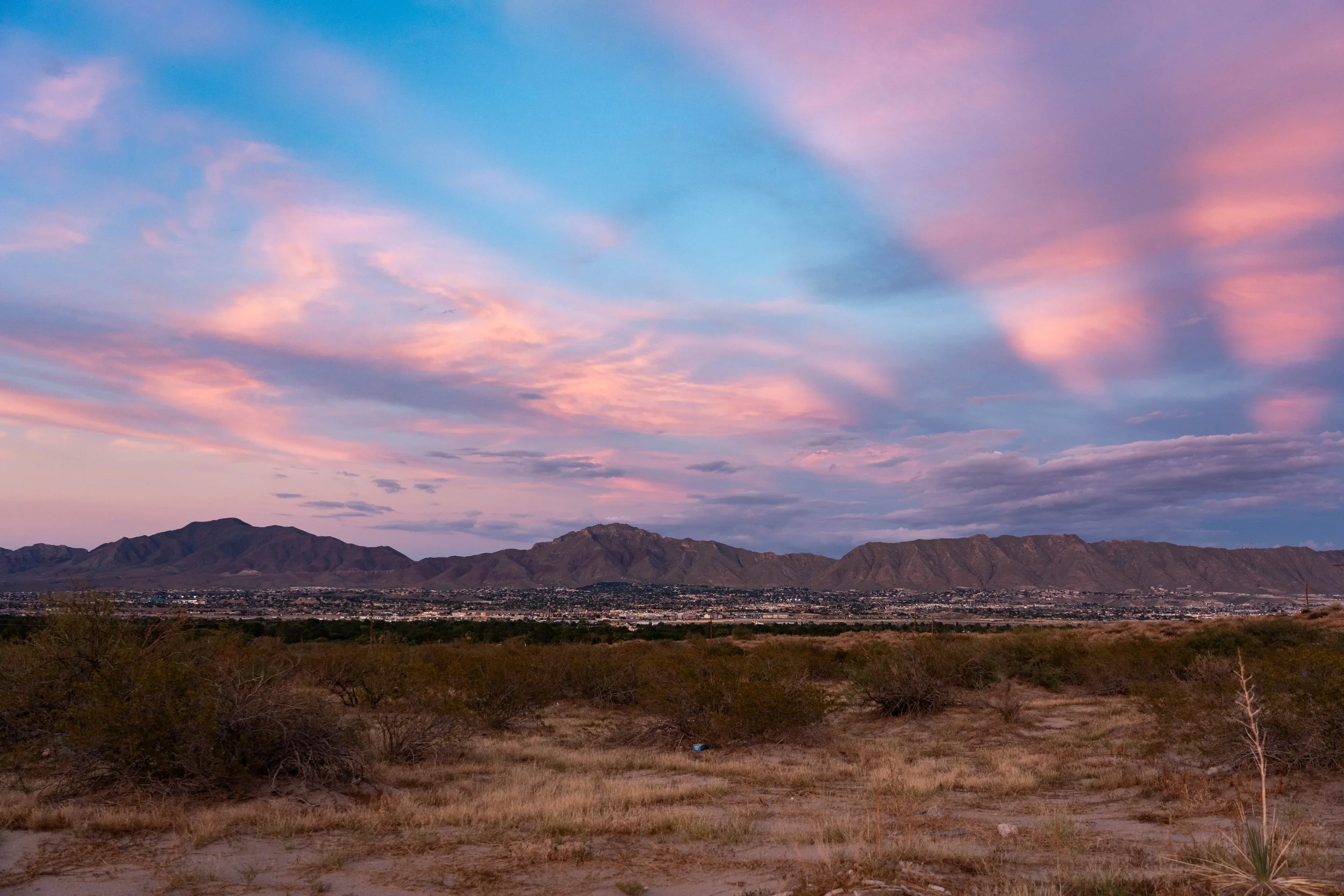 A desert landscape at sunset with mountains in the background and a sky filled with pink, purple, and blue clouds.