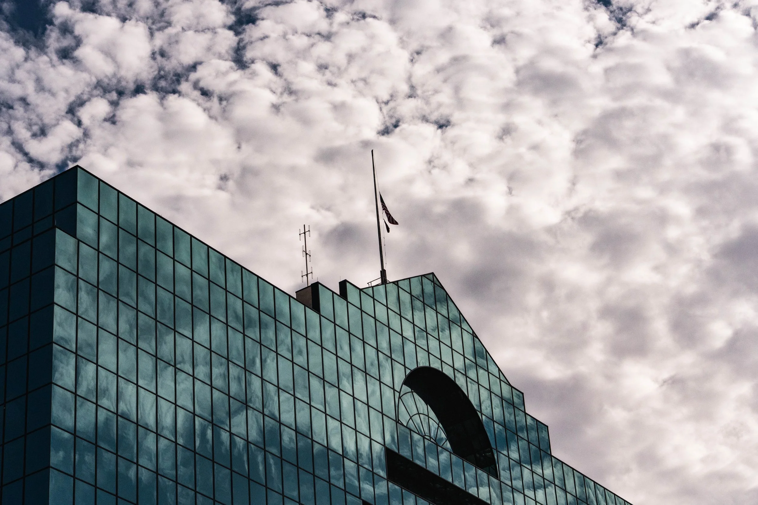 Close-up of a modern glass building with an arched window and flags on top, against a cloudy sky.