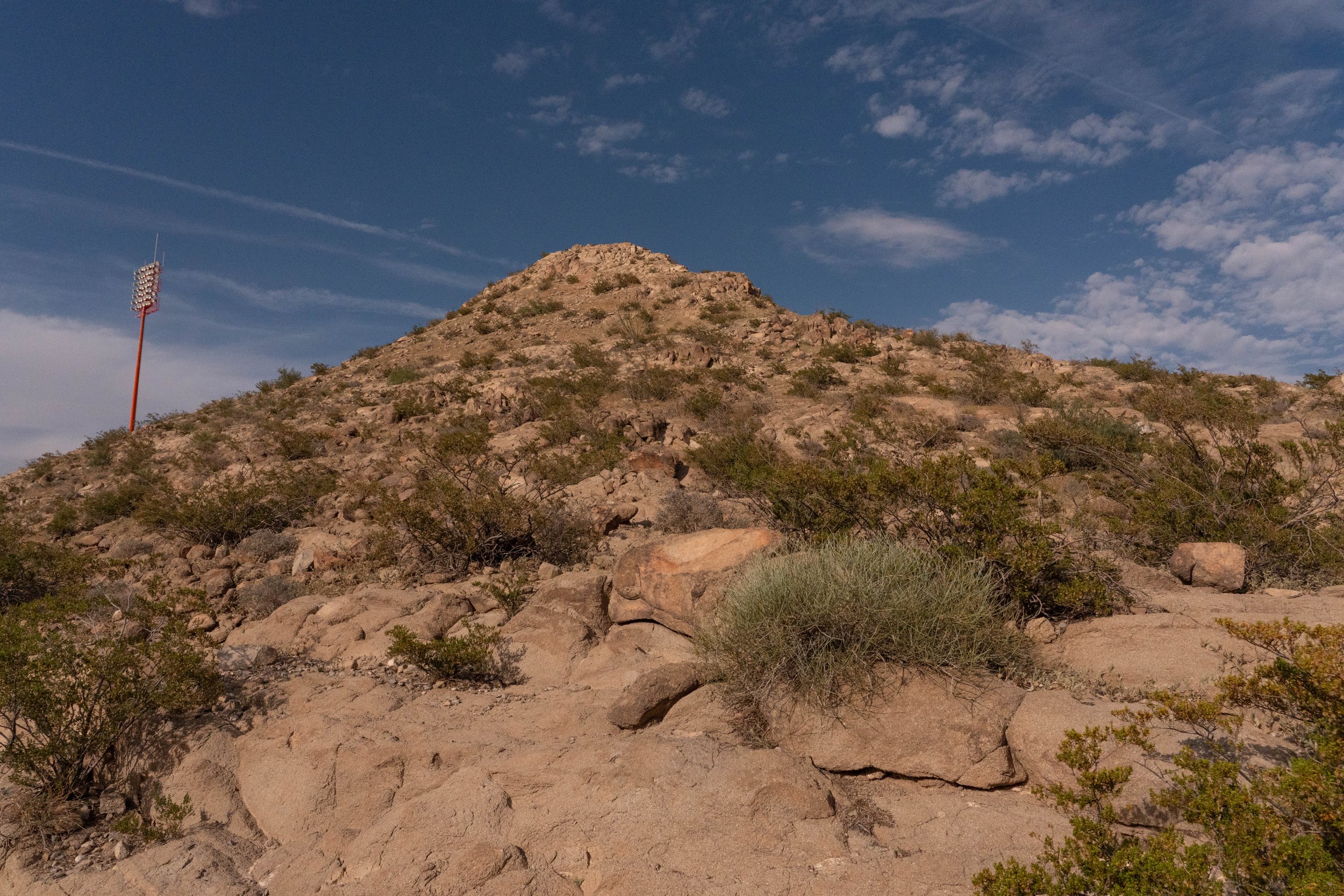 A rocky desert landscape featuring a hillside with sparse desert vegetation and a tall, red stadium light pole on the left. The sky is mostly clear with some scattered clouds.
