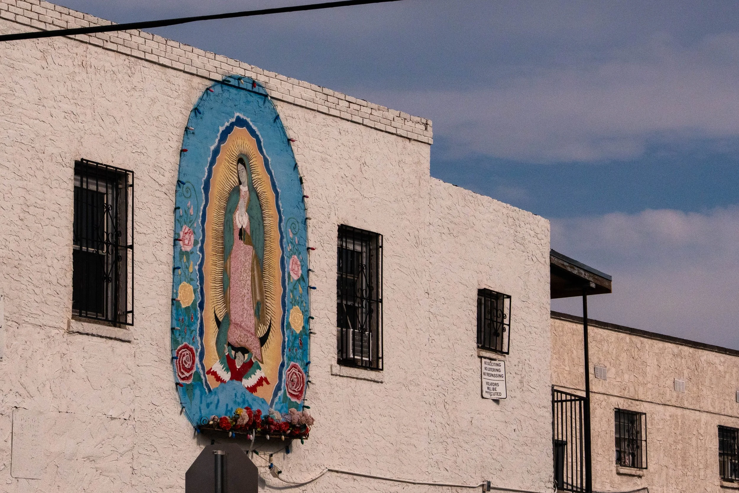 A religious mural depicting Our Lady of Guadalupe on a white stucco building with barred windows, blue sky, and a sign indicating no soliciting, loitering, or trespassing.
