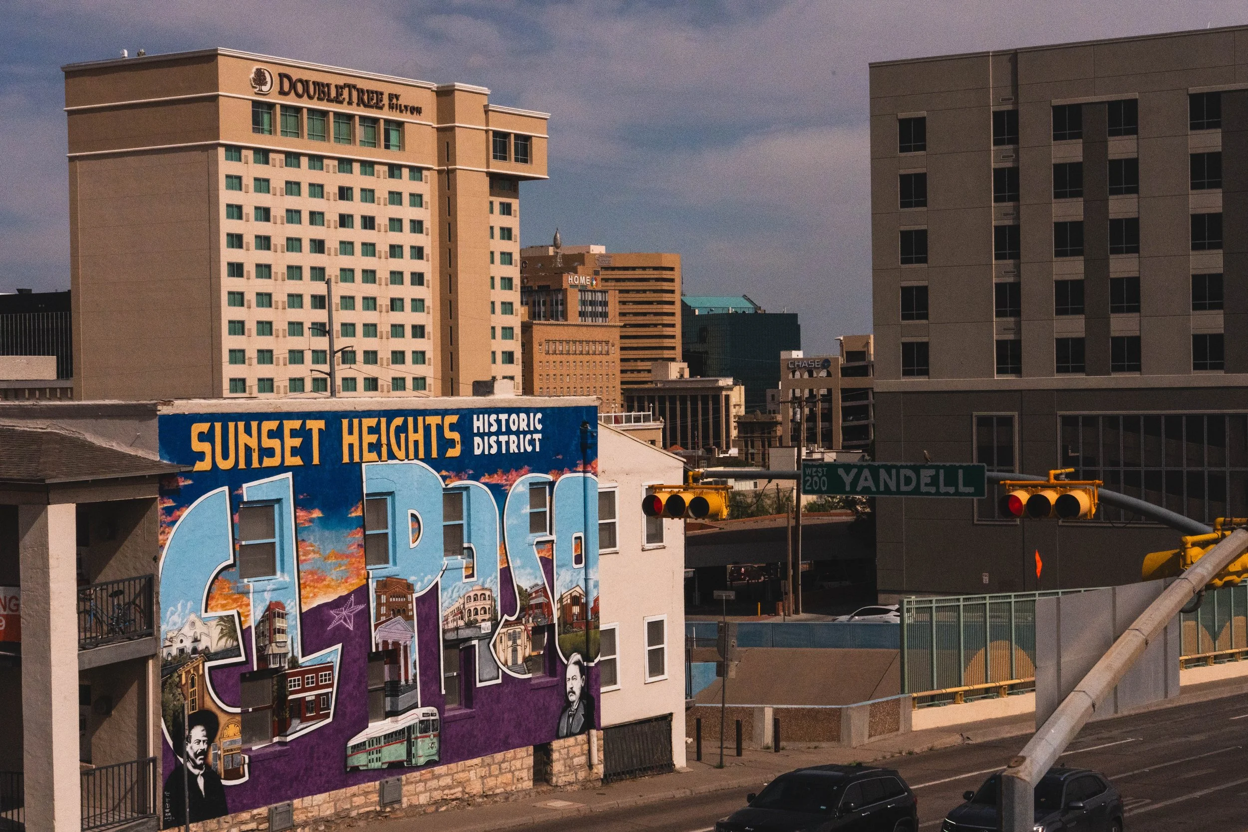 Cityscape featuring buildings and a colorful mural reading 'Sunset Heights Historic District ALICE' with a sunset sky background, street signs, and traffic lights in the foreground.