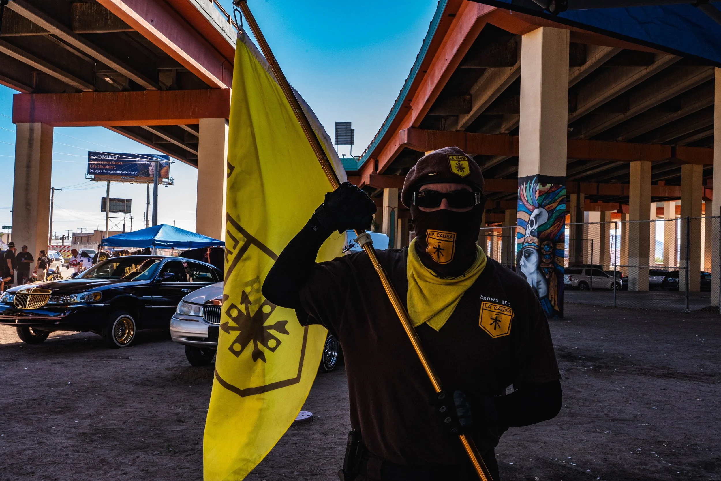 Person holding a yellow flag, dressed in black with a patch that reads 'LA CAUSA,' wearing sunglasses, a face covering, and a badge, standing under an overpass with parked cars and people in the background.