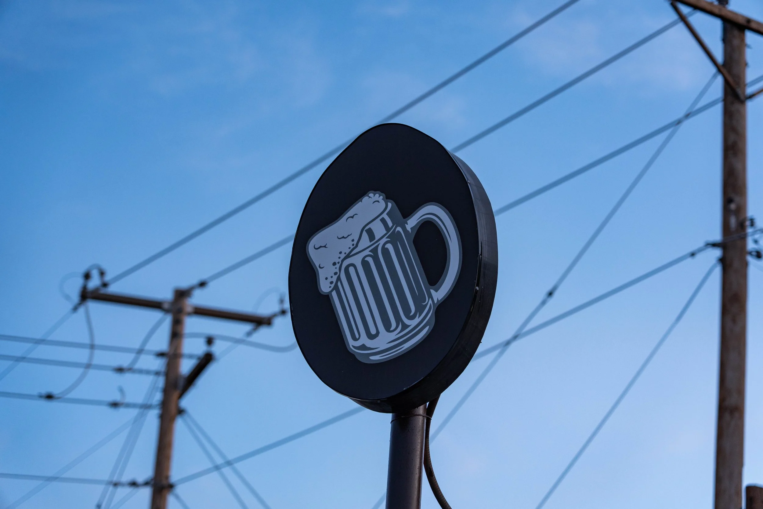 A round sign with a beer mug with foam on top, against a blue sky with power lines and wooden utility poles.