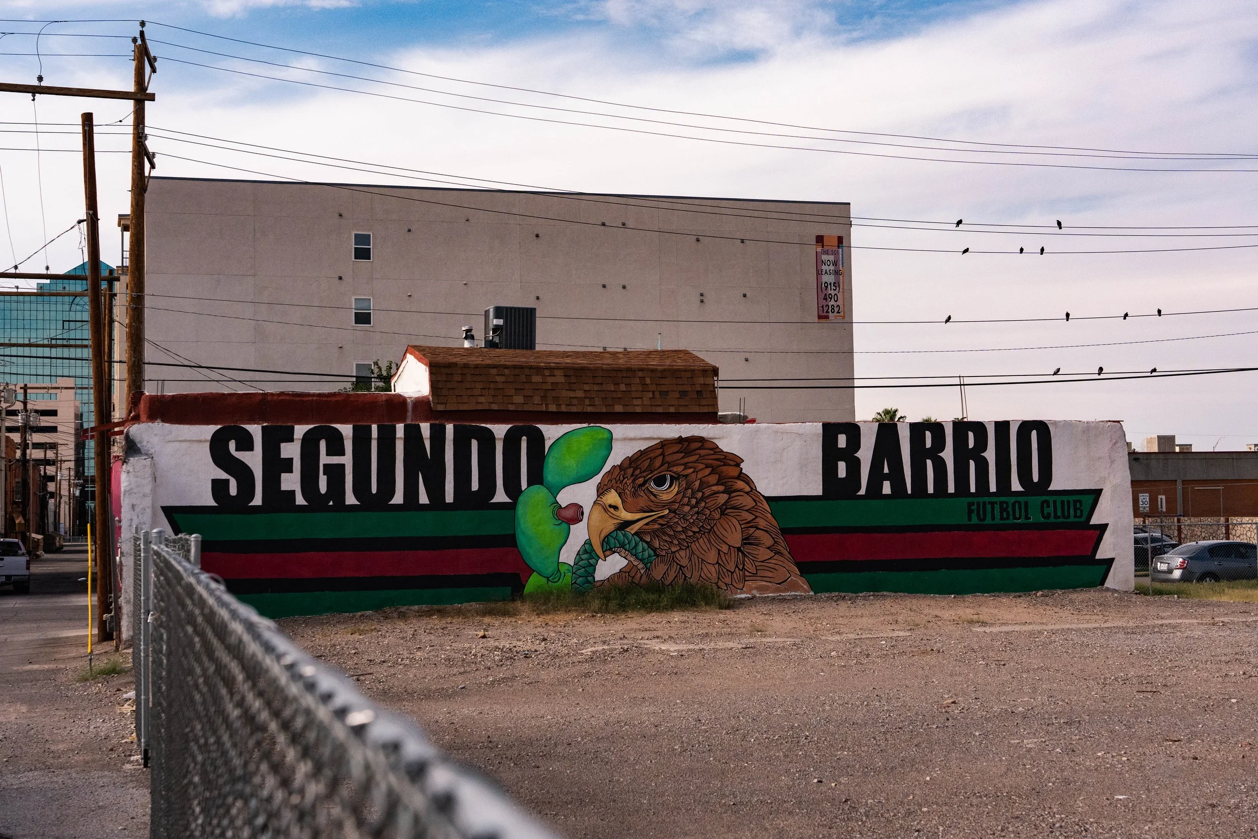 A painted mural on a white wall featuring a bald eagle with a fierce expression clutching a green and brown object, with the words 'Segundo Barrio Futbol Club' in black and green lettering. There is a chain-link fence in the foreground and an urban s