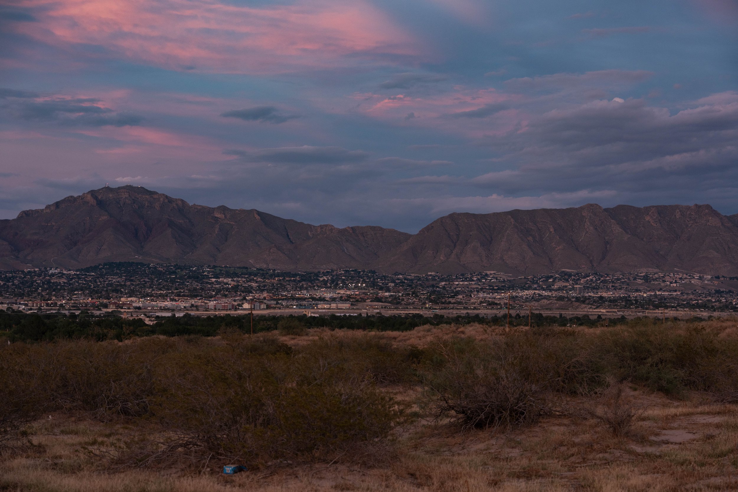 Mountains with a city at their base, under a sky with pink and purple clouds, and desert terrain with bushes in the foreground.