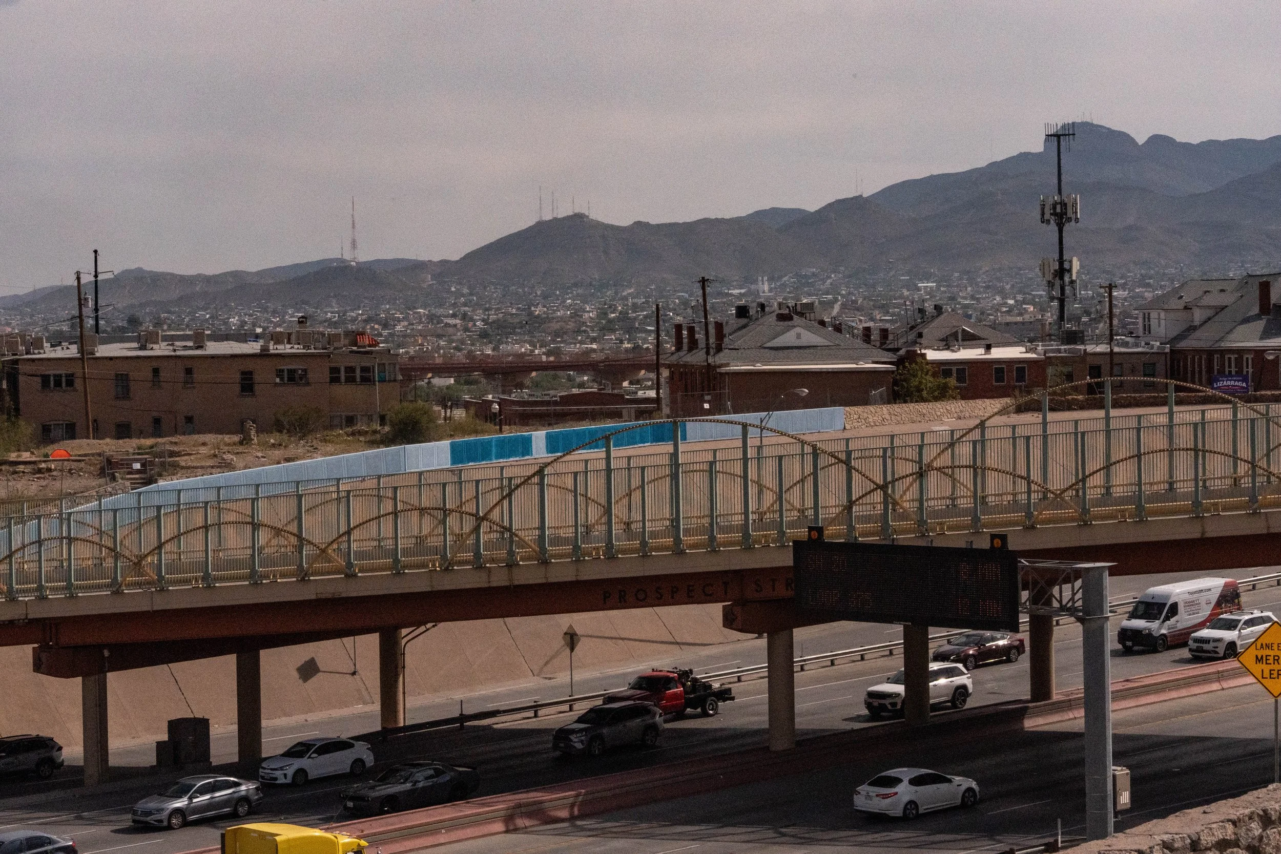 Wide shot of a cityscape with buildings and mountains in the background, viewed from a highway with traffic and a pedestrian bridge.