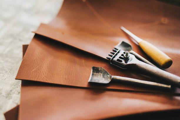 Leatherworking tools on leather pieces, including a utility knife, a squeegee, and a craft knife.